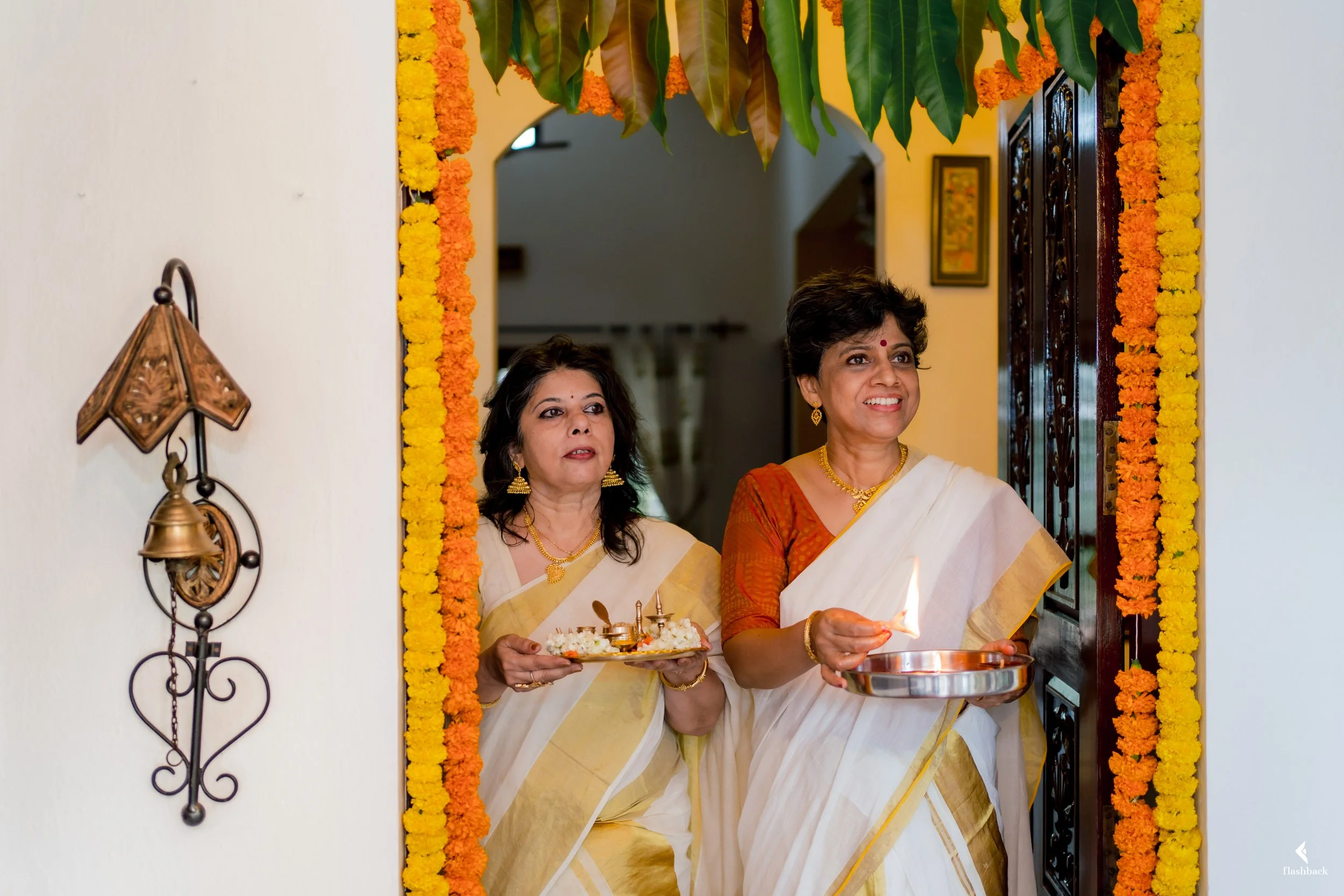 Two women in traditional Indian sarees perform a religious ceremony at the door decorated with marigold flowers, with one woman lighting a flame and the other holding a tray of offerings.