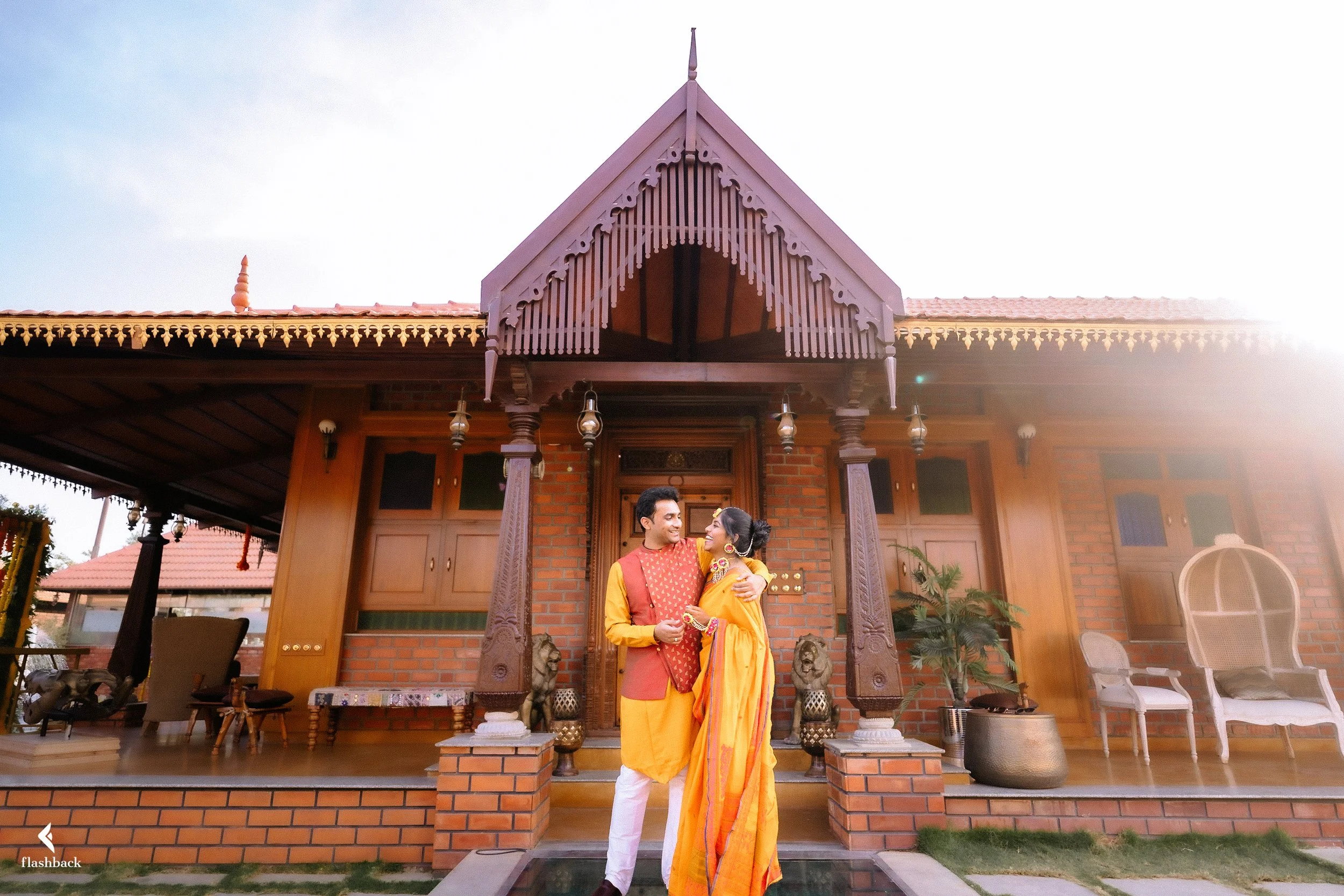 A couple dressed in traditional Indian attire standing outside a brick house with wooden accents, smiling and embracing each other.