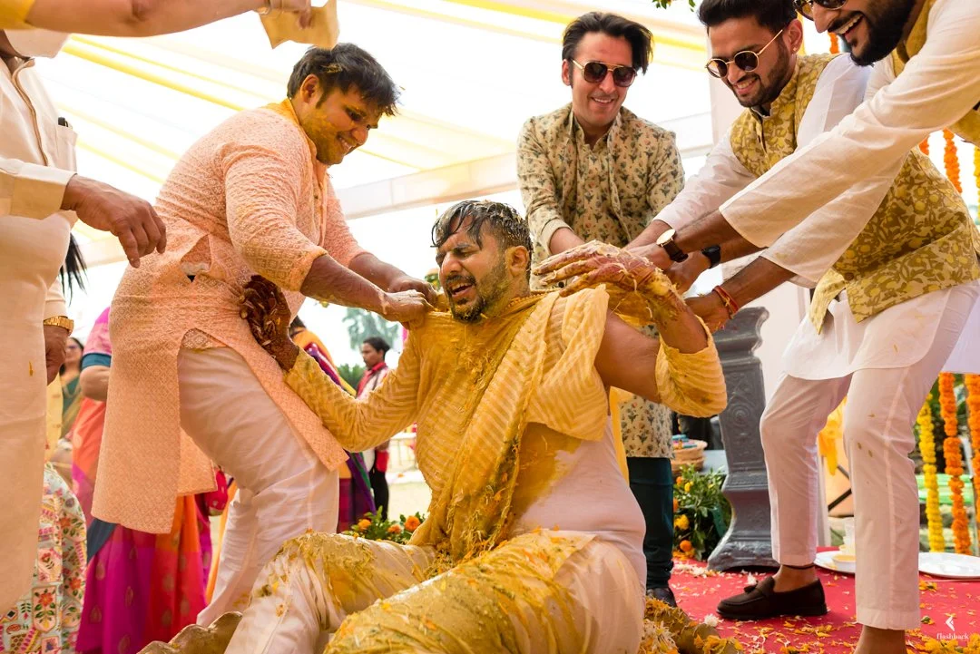 A group of men celebrating a traditional Indian wedding ceremony with a ritual involving yellow paste, with some men smiling and others focused on the ritual, under a decorated canopy.