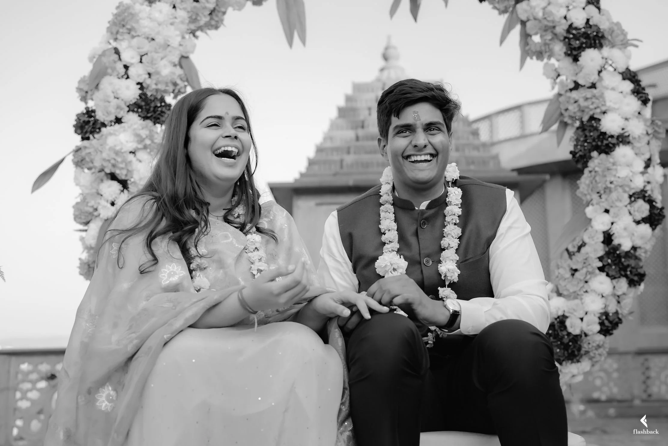 A happy couple dressed in traditional Indian attire sitting together during a celebration, with a floral arch in the background.