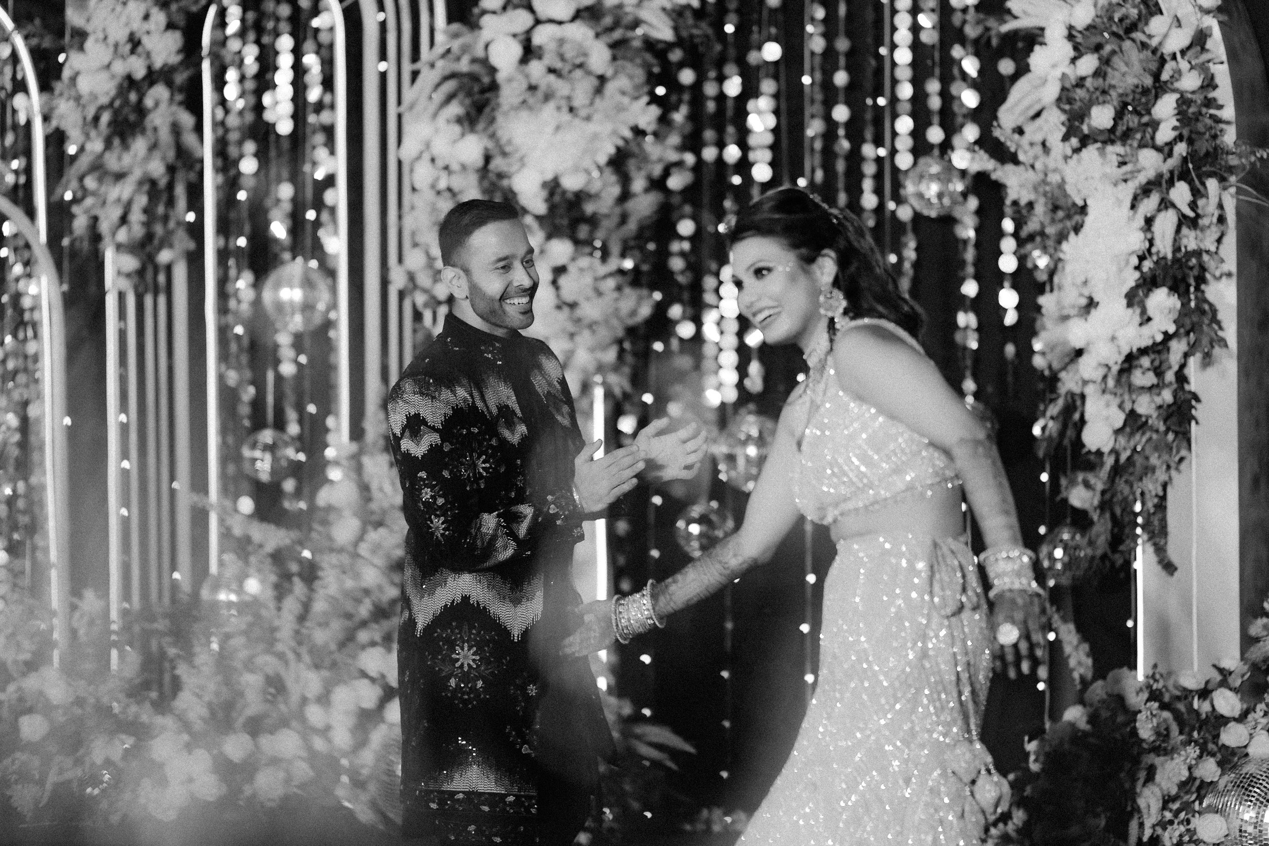 A couple dancing at a wedding reception, with the man wearing a kimono-style jacket and the woman in a sparkling traditional dress, amidst floral and decorative lights.