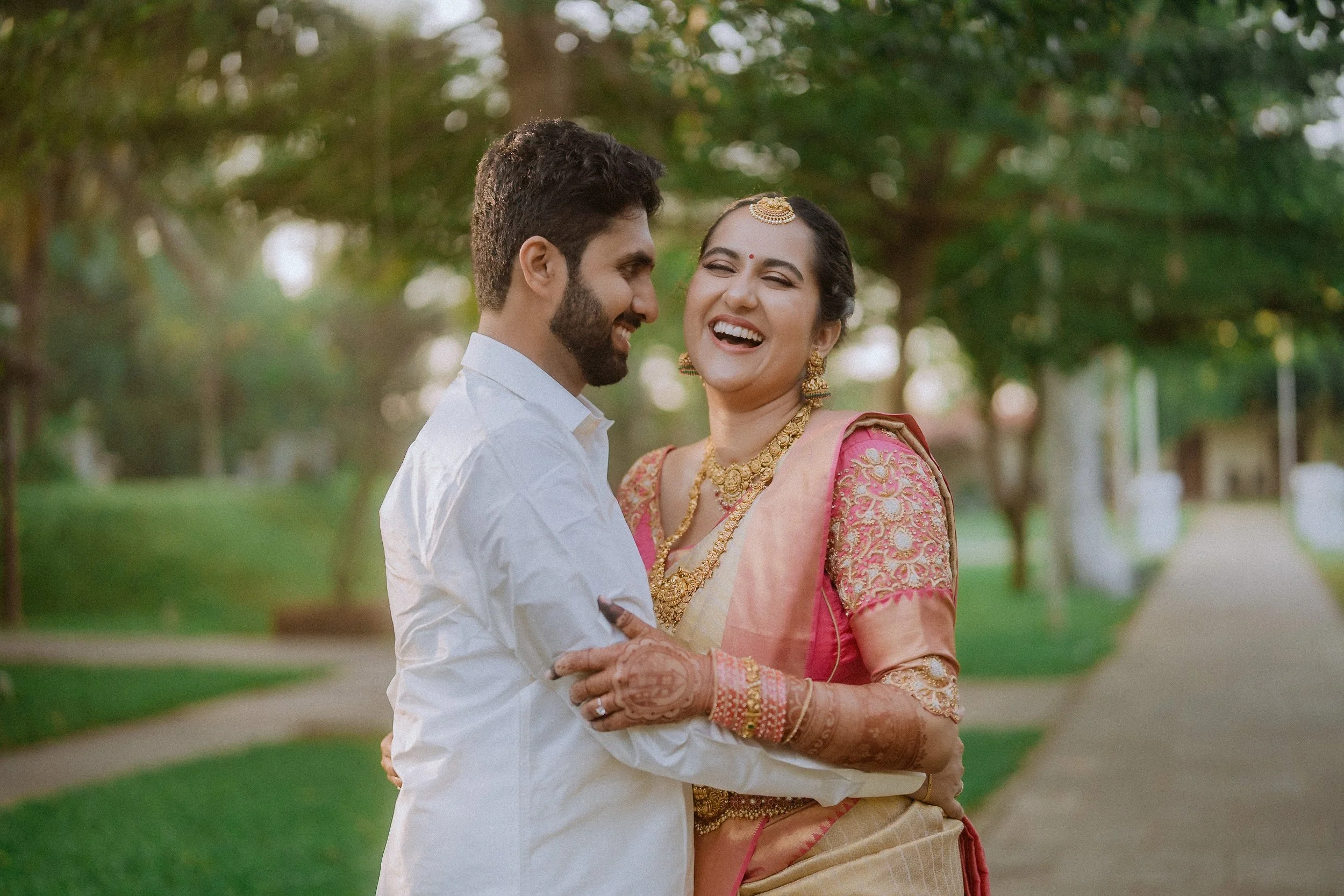 A man and woman share a joyful moment outdoors, smiling and embracing each other. The woman is dressed in traditional Indian attire with jewelry, and the man is wearing a white shirt. Trees and a pathway are visible in the background.