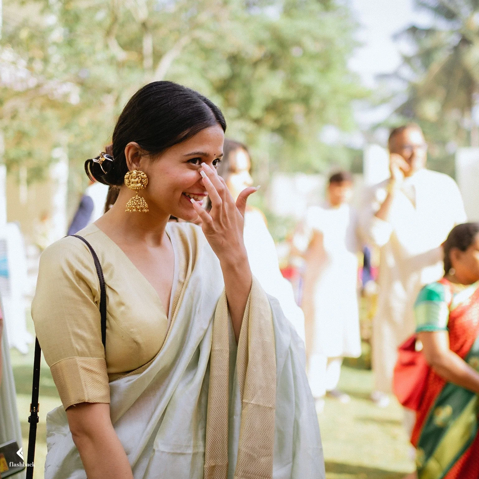 A woman in a cream sari laughing and touching her nose at an outdoor event, surrounded by other people, some dressed in traditional Indian attire.
