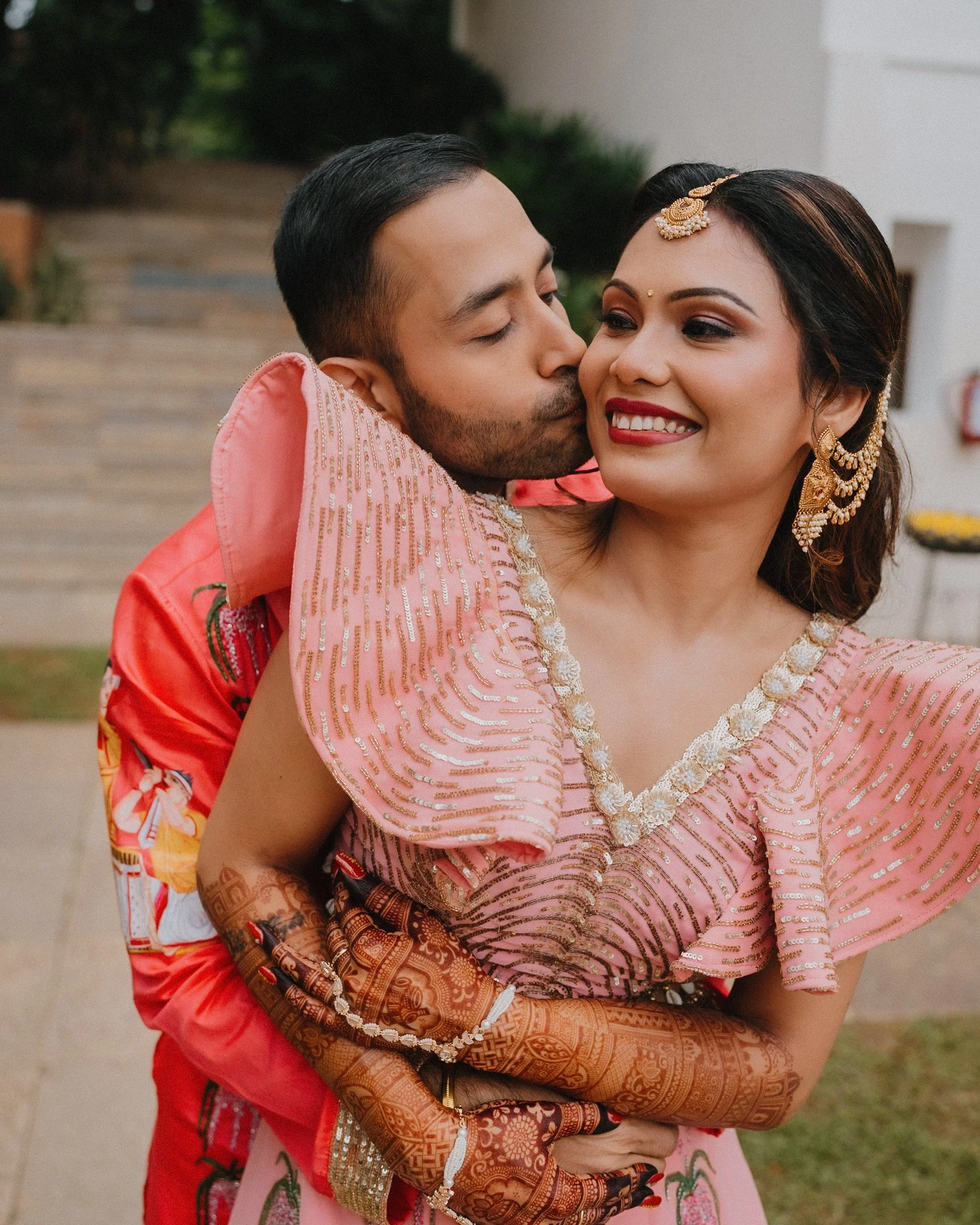 A couple dressed in traditional Indian attire embracing outdoors, with the man kissing the woman on her cheek. The woman is smiling, wearing intricate jewelry, eye makeup, and a pink outfit with gold embellishments. The man is wearing a red outfit, a
