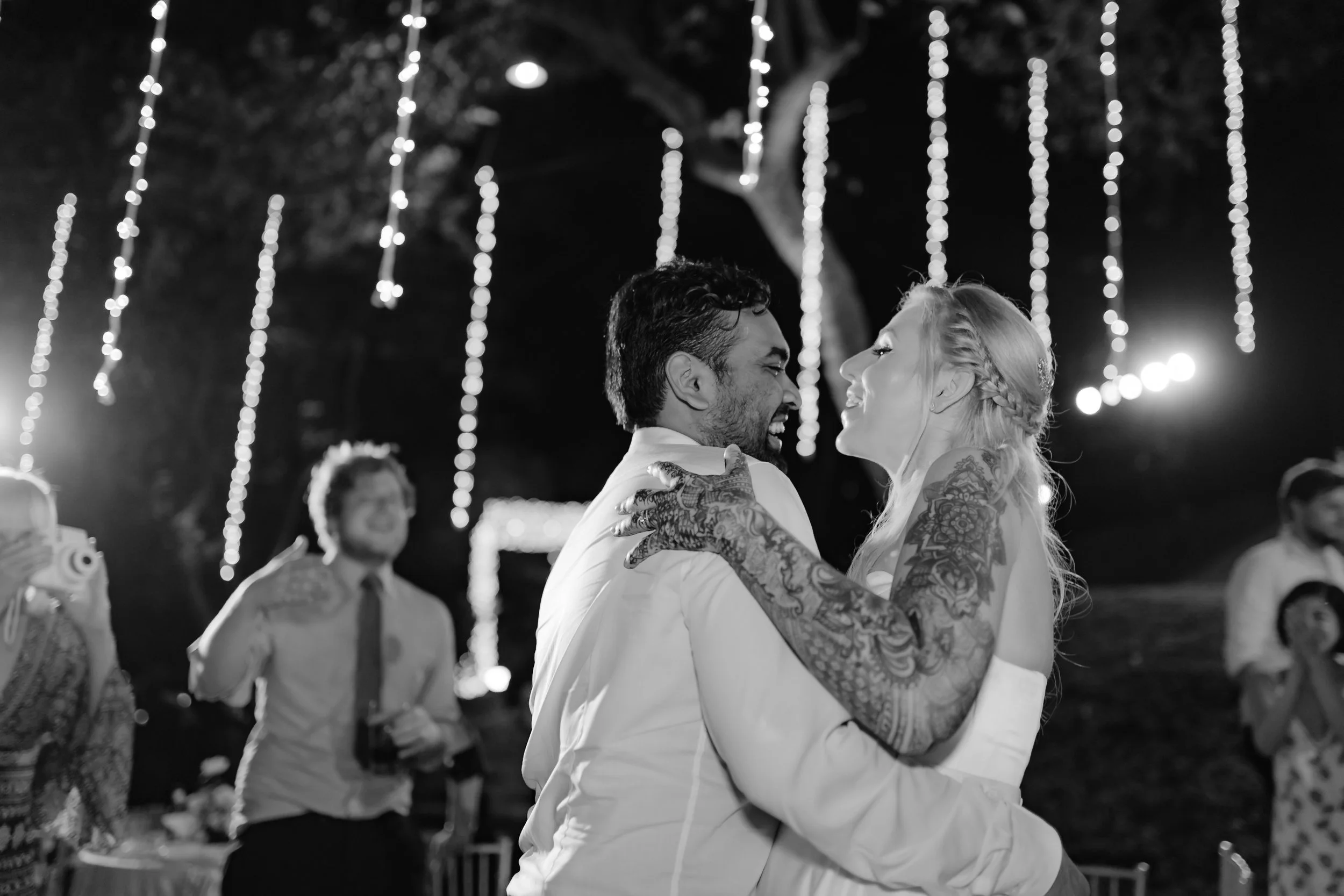 A black and white photo of a couple dancing closely at night, surrounded by string lights, with guests in the background.