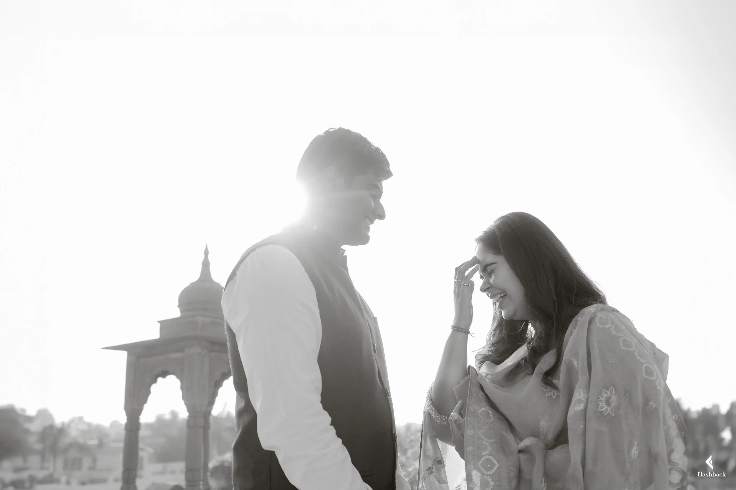 A man and woman smiling at each other outdoors, backlit by the sun, with a historic monument in the background