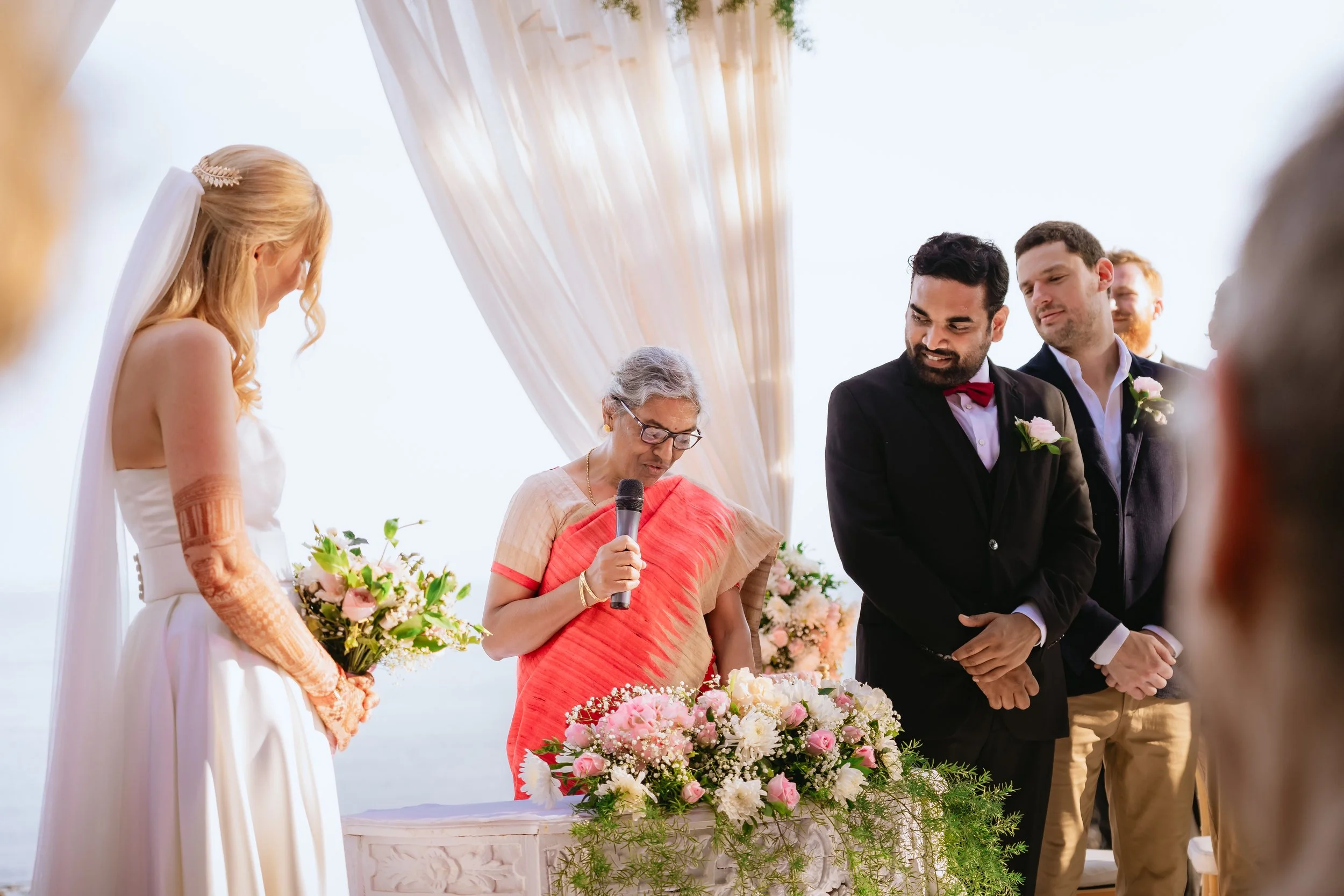 A wedding ceremony with a bride, groom, officiant, and guests outdoors decorated with flowers and a draped canopy.