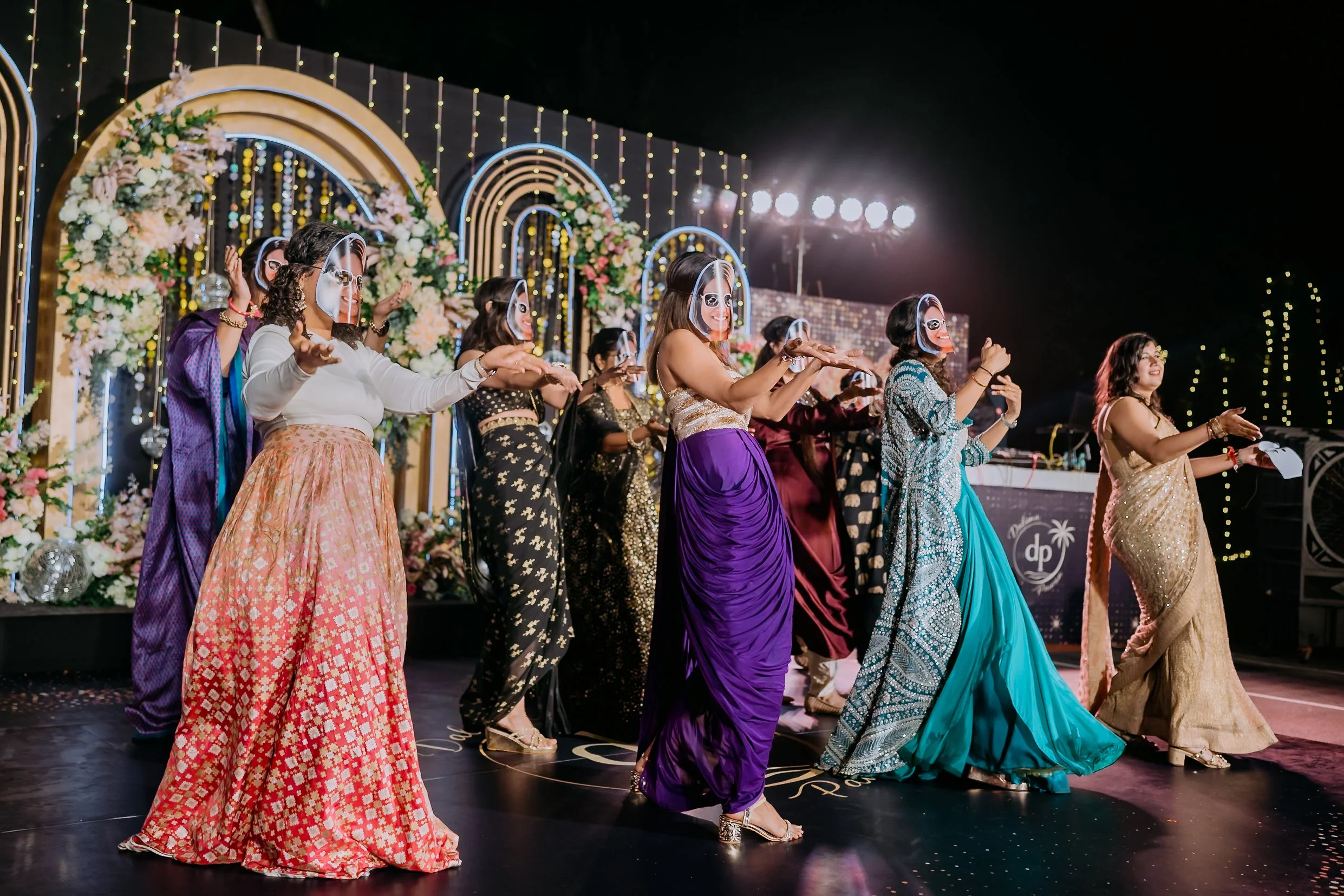 A group of women at a festive event, wearing colorful sarees, dancing and smiling under decorative lights and floral arrangements.