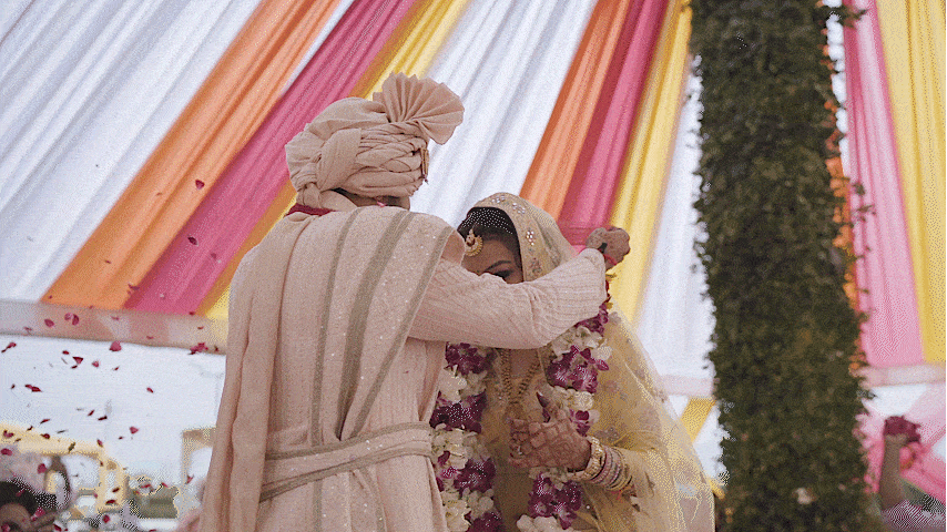 A traditional Indian wedding ceremony with the groom adjusting the bride's head covering in a decorated outdoor tent with colorful drapes and floral decorations.