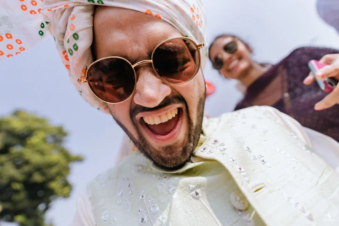A man wearing sunglasses, a patterned headscarf, and a cream-colored traditional outfit smiling and looking at the camera, with a woman in sunglasses and a dark outfit smiling in the background.