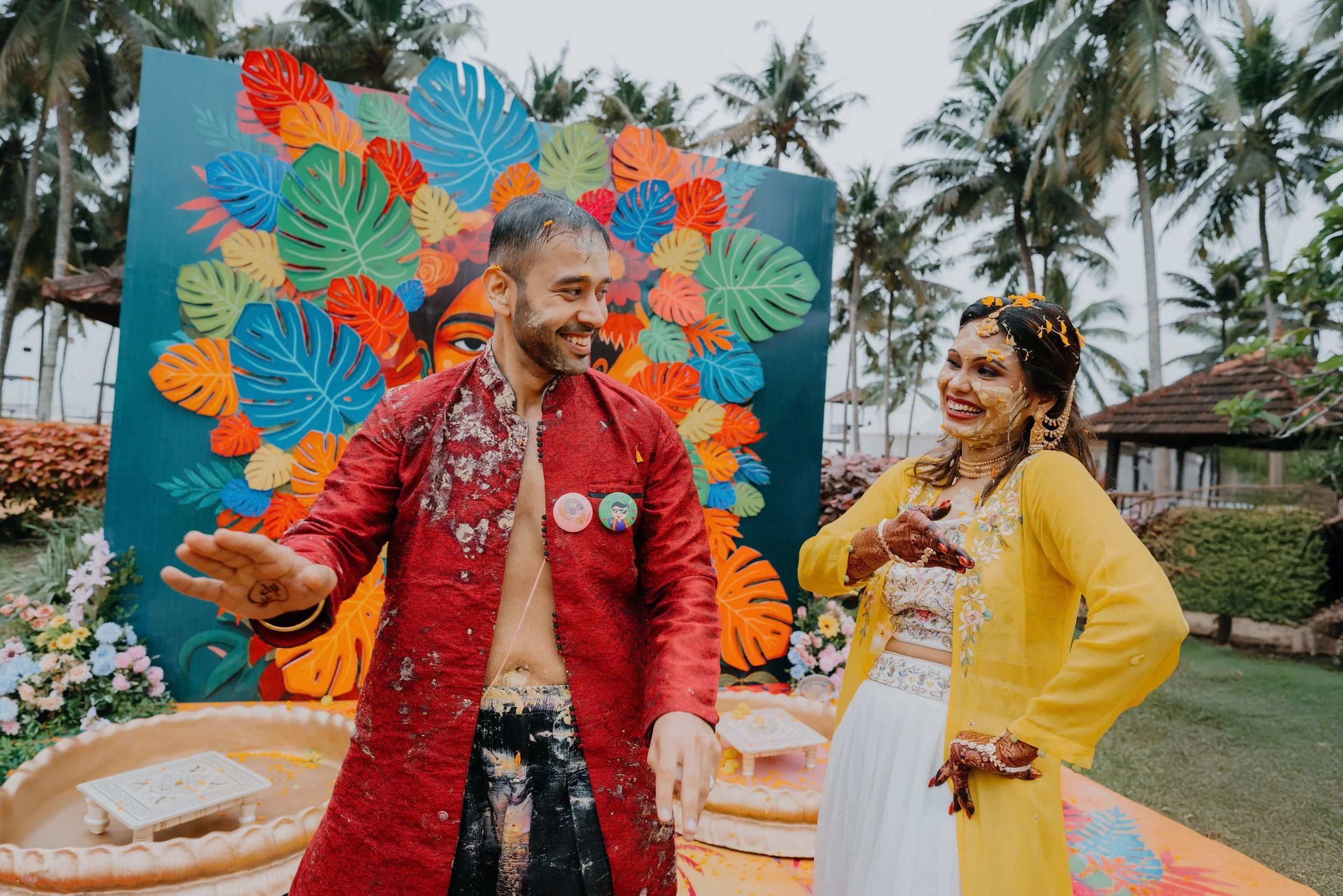 A man and woman celebrating at a colorful outdoor event with tropical-themed decorations, including large leaves and vibrant patterns, while the man throws flower petals and the woman smiles with flower petals on her face.