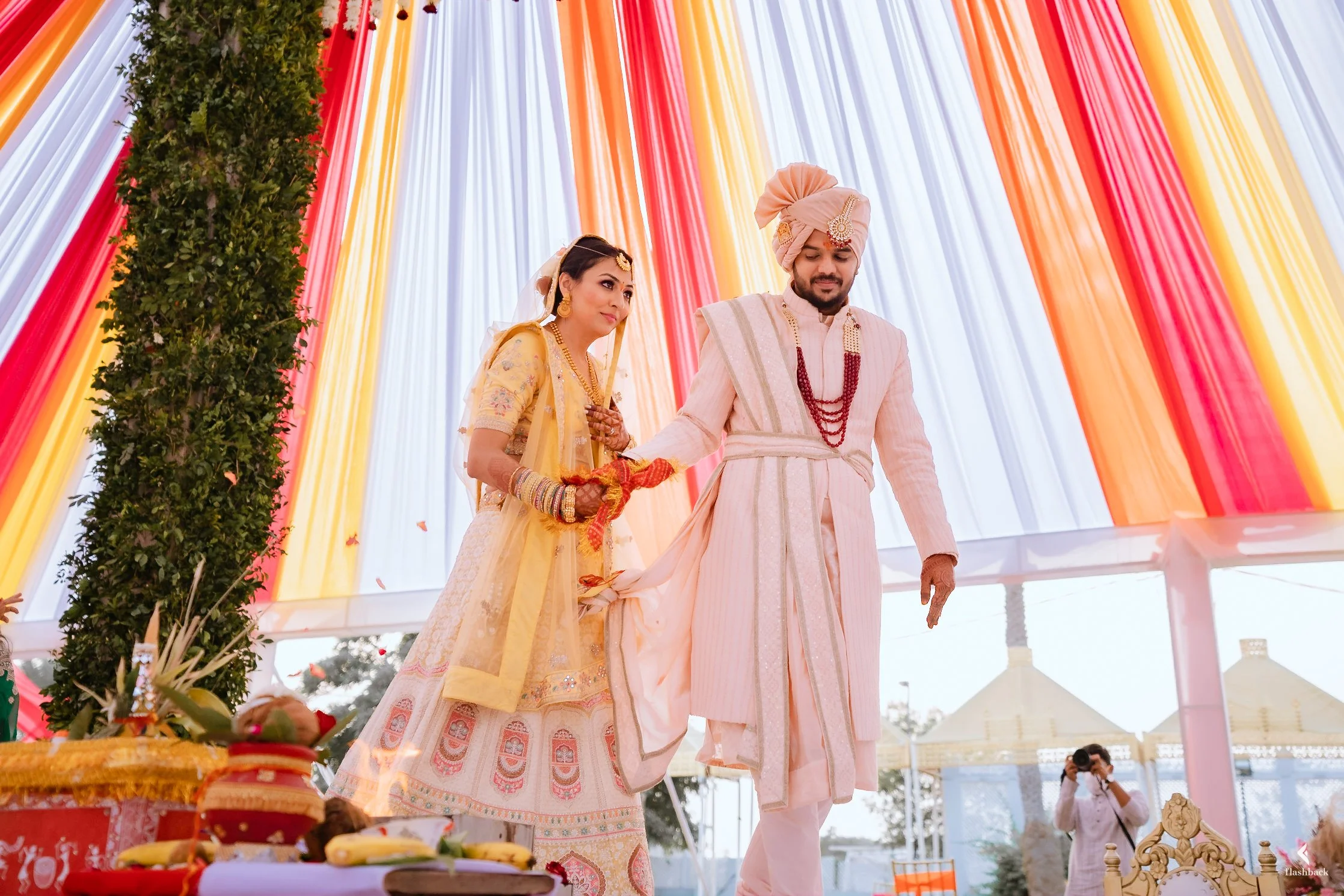 Indian bride and groom performing a wedding ritual under a colorful decorated tent.