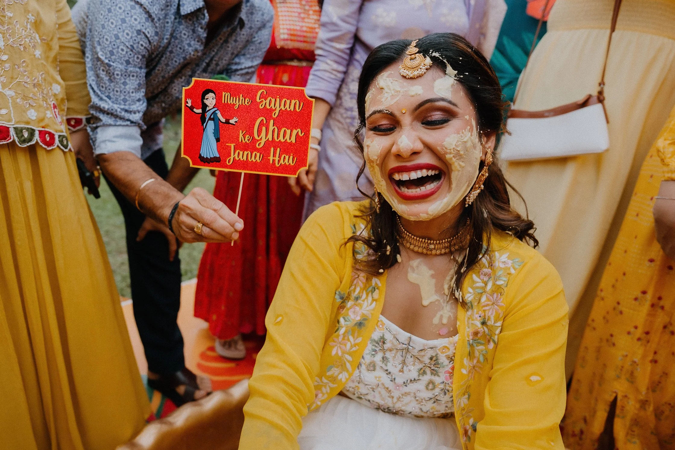 A woman with dark hair and traditional jewelry is laughing with her eyes closed. She is covered in a yellow garment, and others around her are holding a colorful sign with a cartoon woman and Hindi text that reads "Mayhe Sajan ke Ghar Jan Hai." The b
