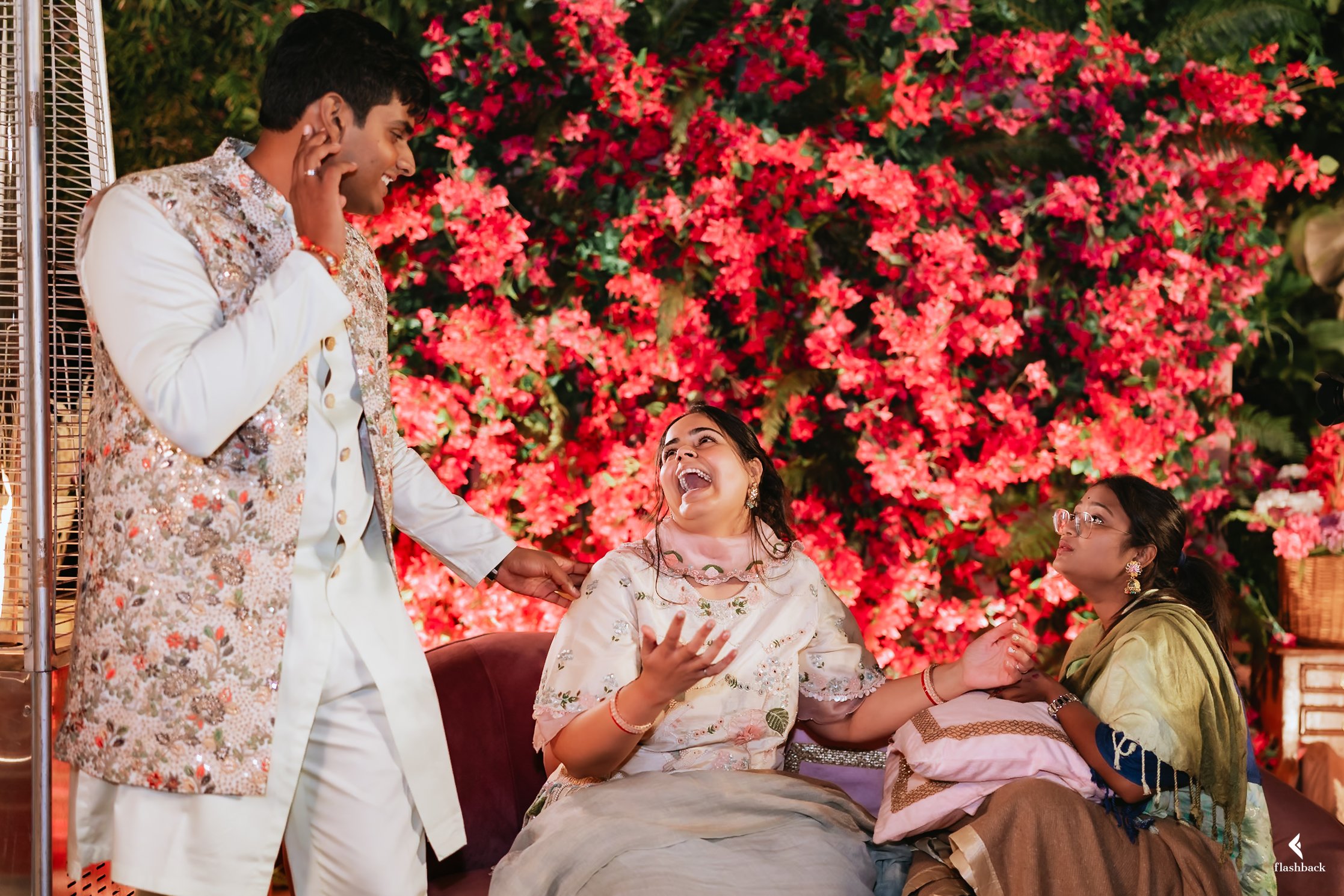 A joyful Indian wedding celebration with four people; a man in a white sherwani with embroidered details, a woman in a cream-colored dress laughing, another woman in a yellow-green saree with glasses, all seated or standing against a backdrop of vibr
