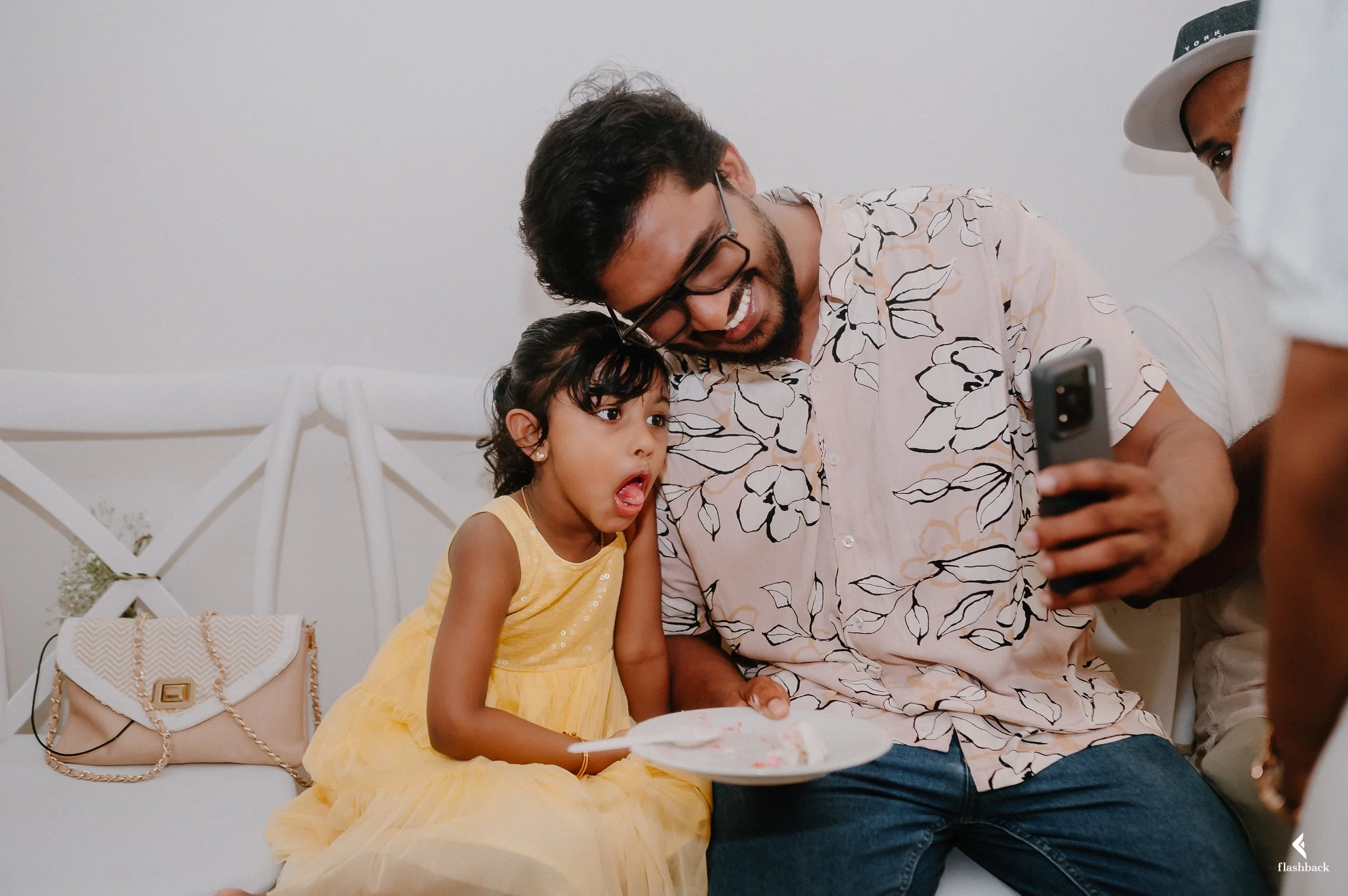 A man, a young girl, and a person taking a selfie, sitting on a white couch. The girl is wearing a yellow dress, looking surprised, holding a plate with cake. The man is smiling, wearing glasses and a floral shirt. The person taking the photo is part