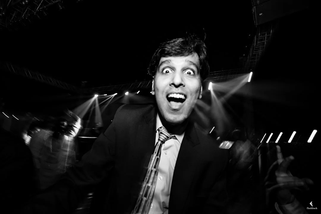 A man with a big smile and wide eyes, wearing a suit and striped tie, at a lively party or club with bright stage lights and blurred background.