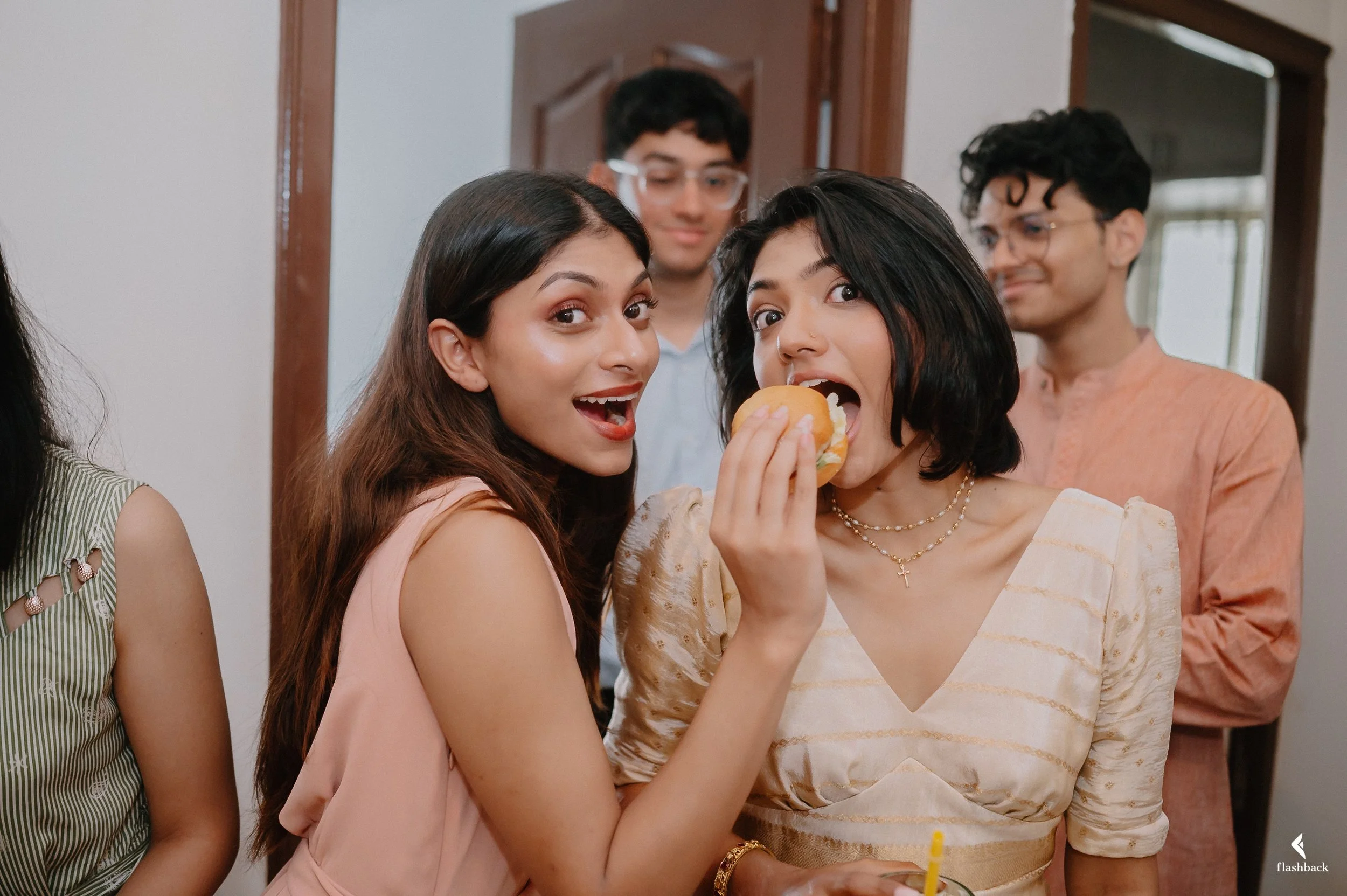Two women smiling and one woman biting into a hamburger at a gathering, with two men in the background.