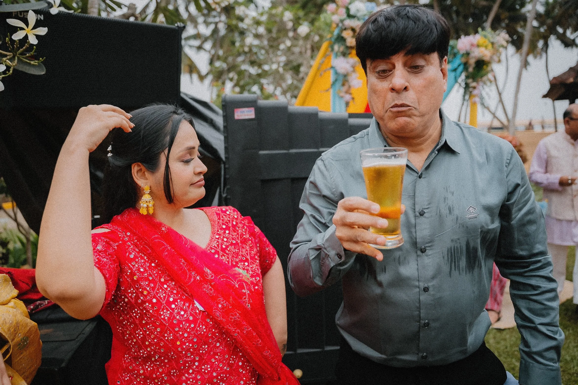 A woman in a red traditional dress with earrings sitting at a table, and a man in a gray shirt holding a glass of beer, both at an outdoor celebration with floral decorations and trees in the background.