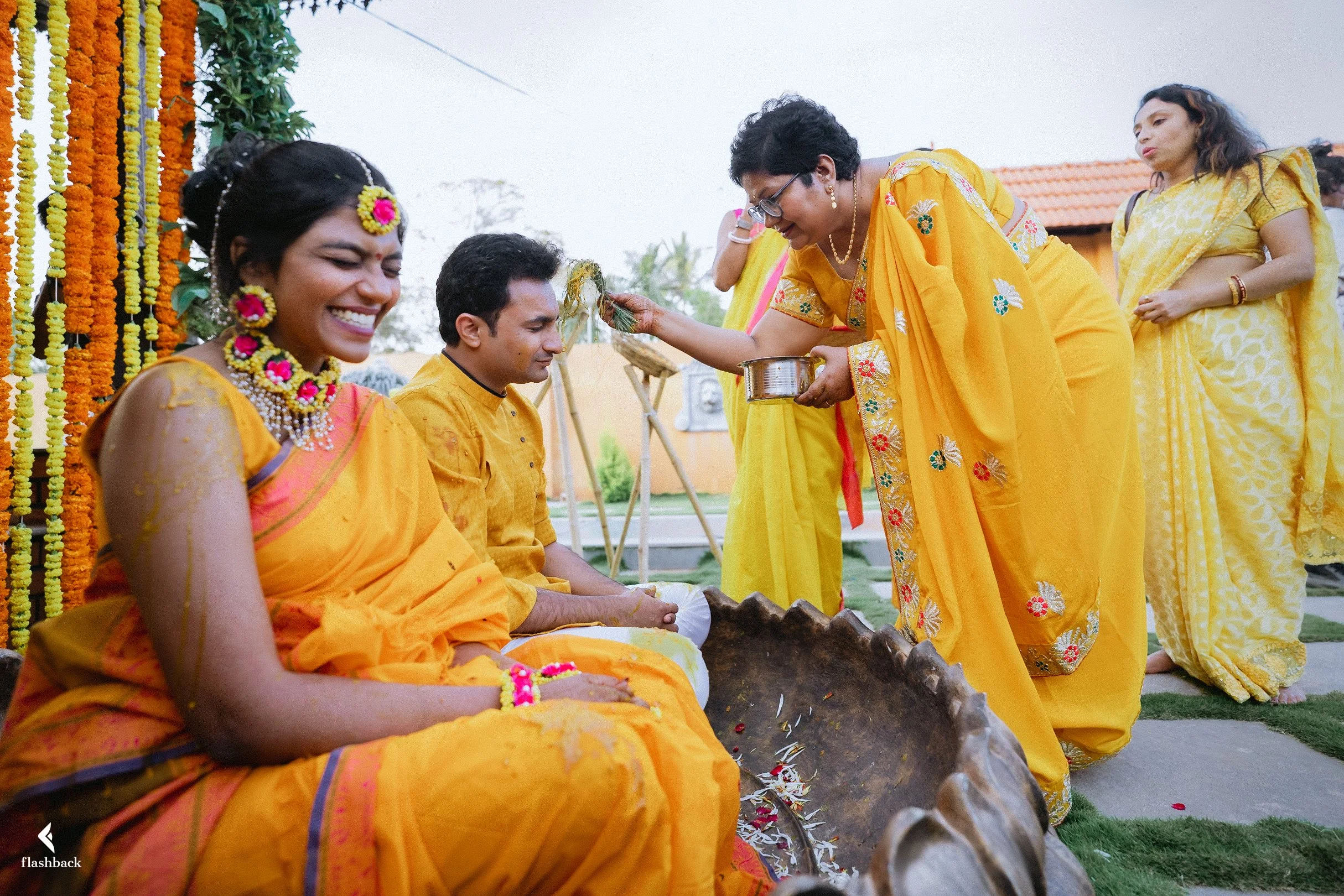 A traditional Hindu wedding ceremony with women dressed in yellow sarees, some with jewelry, participating in rituals outdoors on a decorated platform with orange marigold garlands.