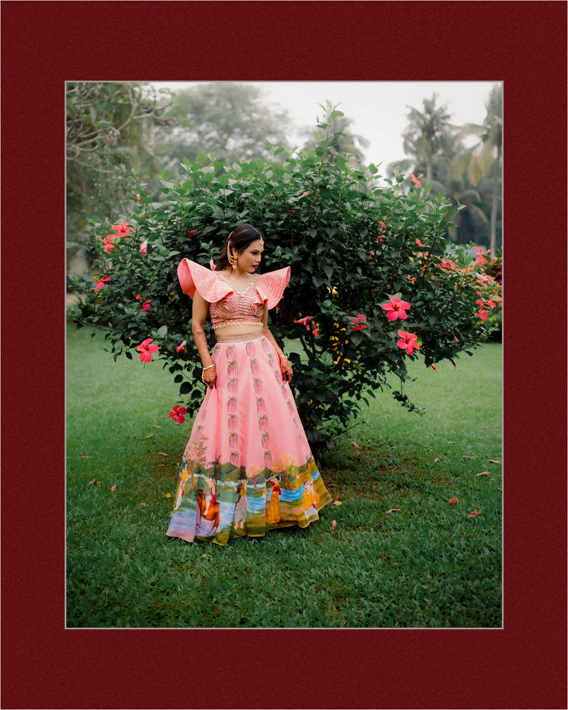 A woman in traditional attire standing outdoors near pink hibiscus flowers and green foliage.