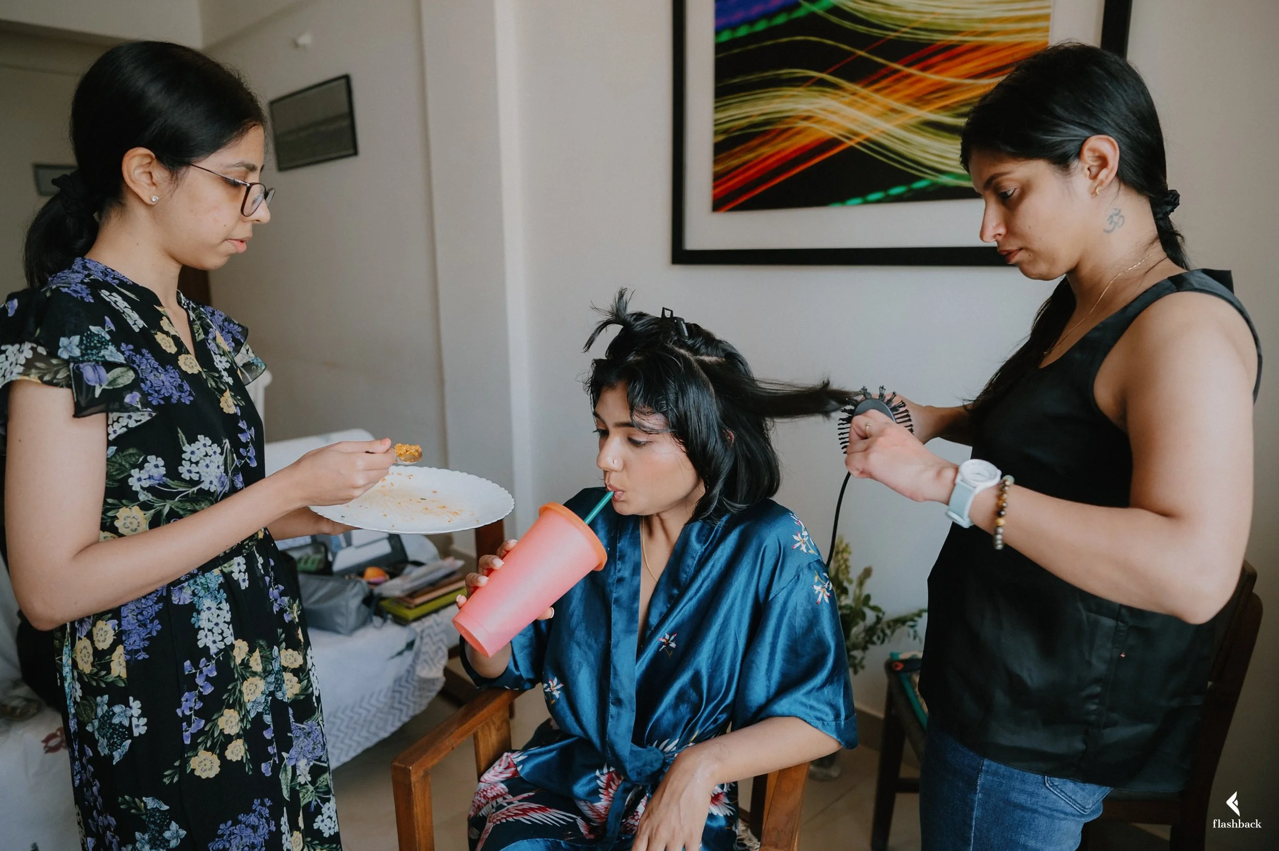 Three women in a room, one sitting in a chair drinking from a pink cup with a straw, another woman styling her hair with a curling iron, and a third woman holding a plate of food.