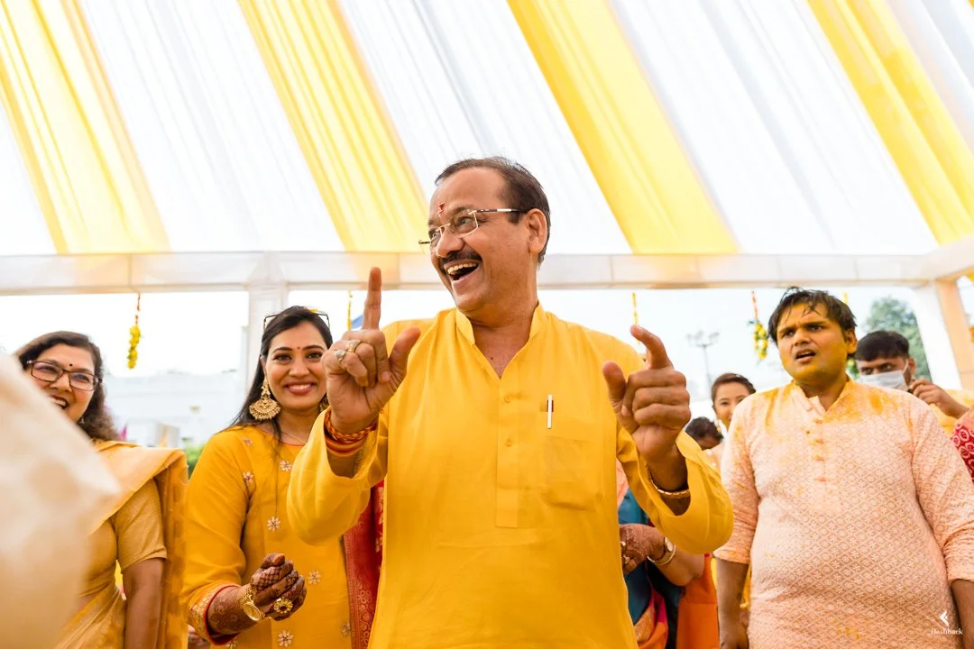A group of people celebrating at a gathering, with a man in a yellow kurta smiling and gesturing with his hands.