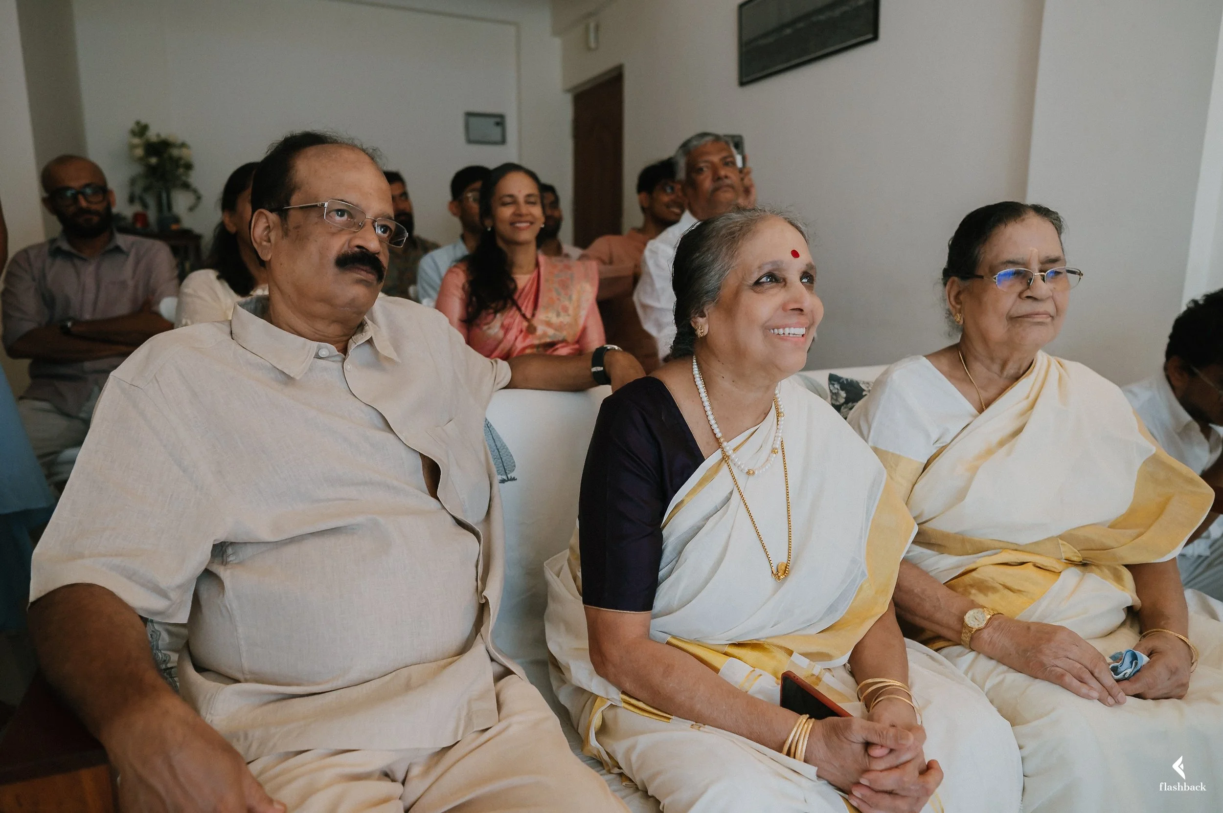 Group of Indian people attending a formal event, sitting and standing, with some smiling and wearing traditional Indian sarees and kurtas.