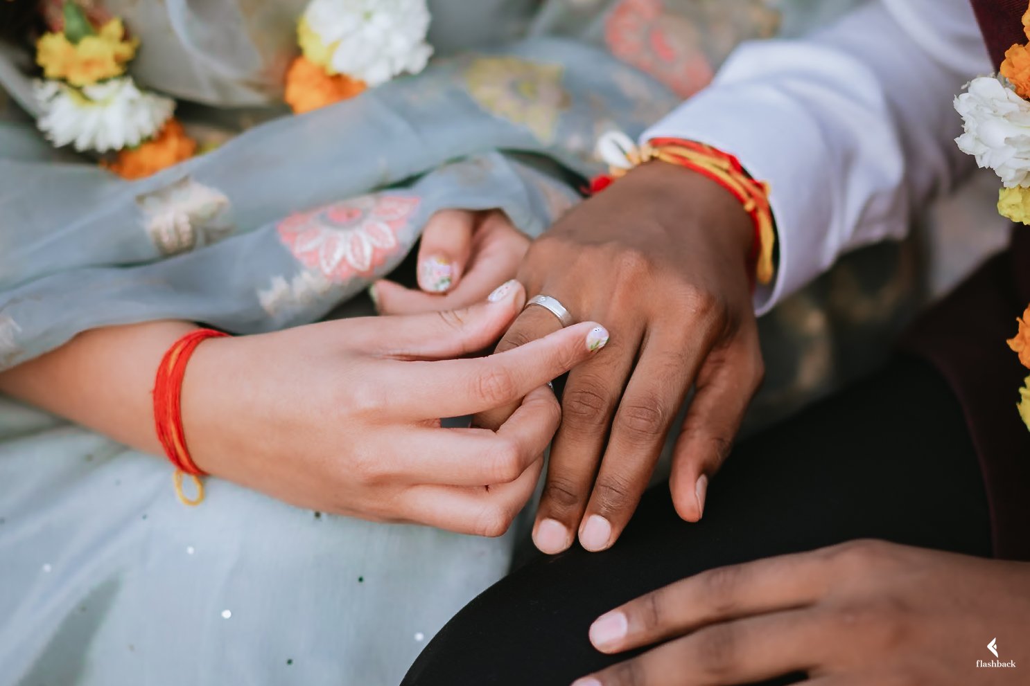 Close-up of a person's hand holding another person's hand gently during a celebration, with both wearing colorful bracelets and traditional attire, surrounded by flower garlands.