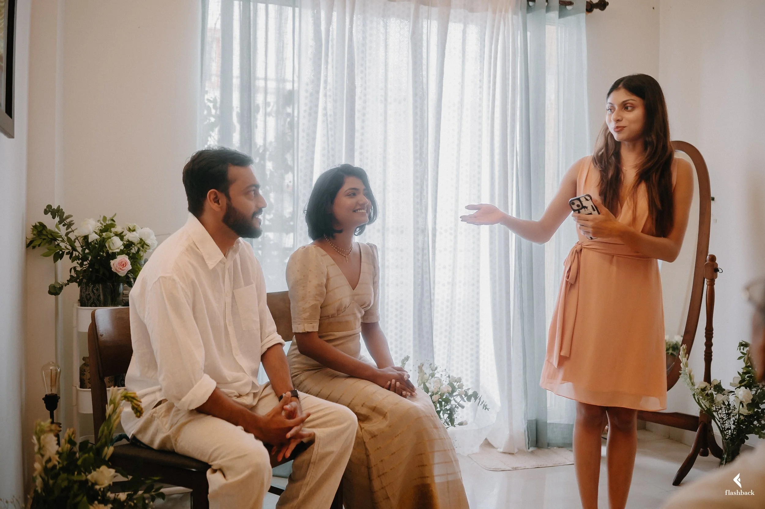A woman stands holding a phone and gesturing towards two people seated on a chair, smiling and engaged in conversation in a bright room with sheer curtains, flowers, and a wooden chair.