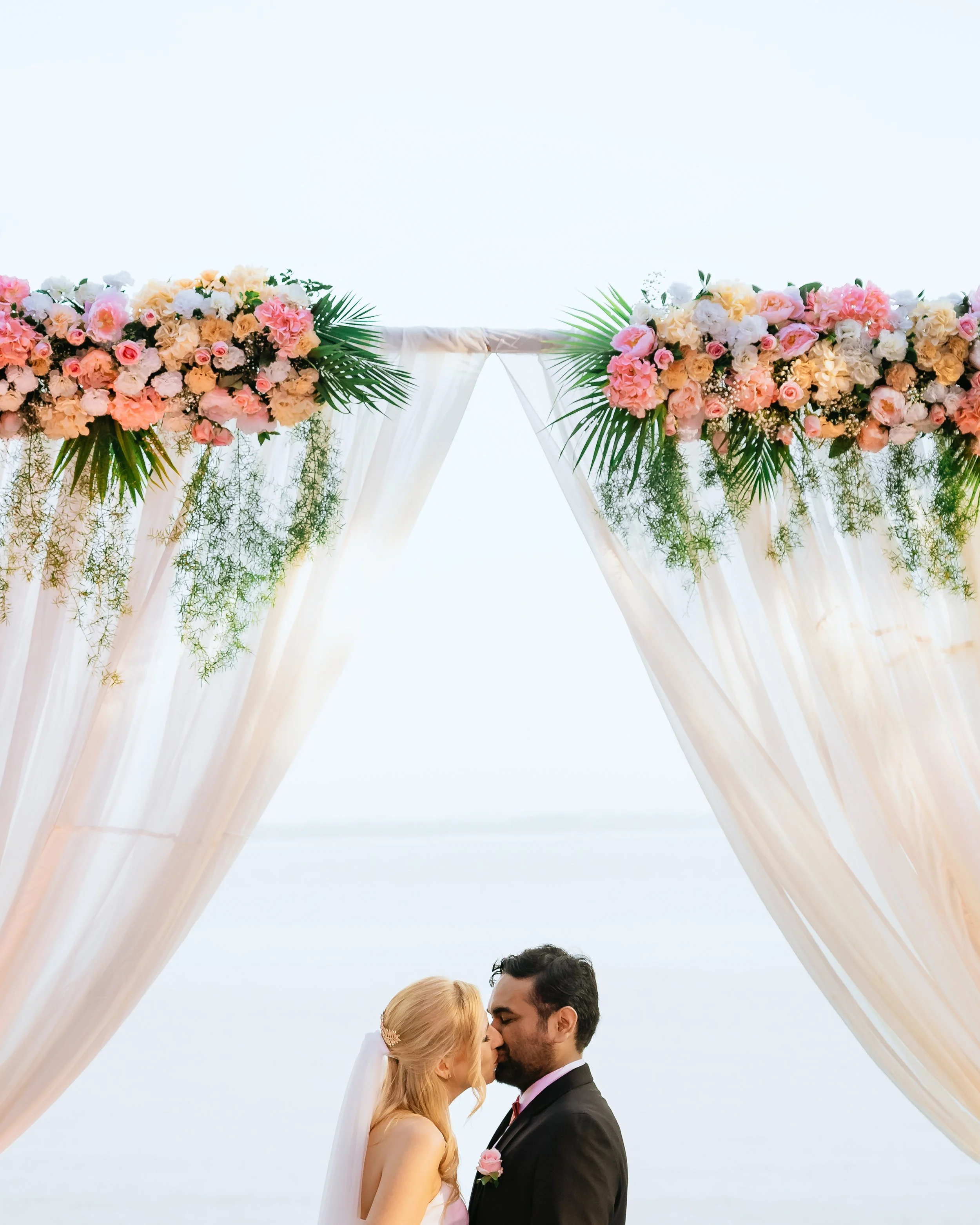 A bride and groom sharing a kiss under a floral wedding arch with pink, white, and peach roses, and green foliage, set outdoors by a body of water.
