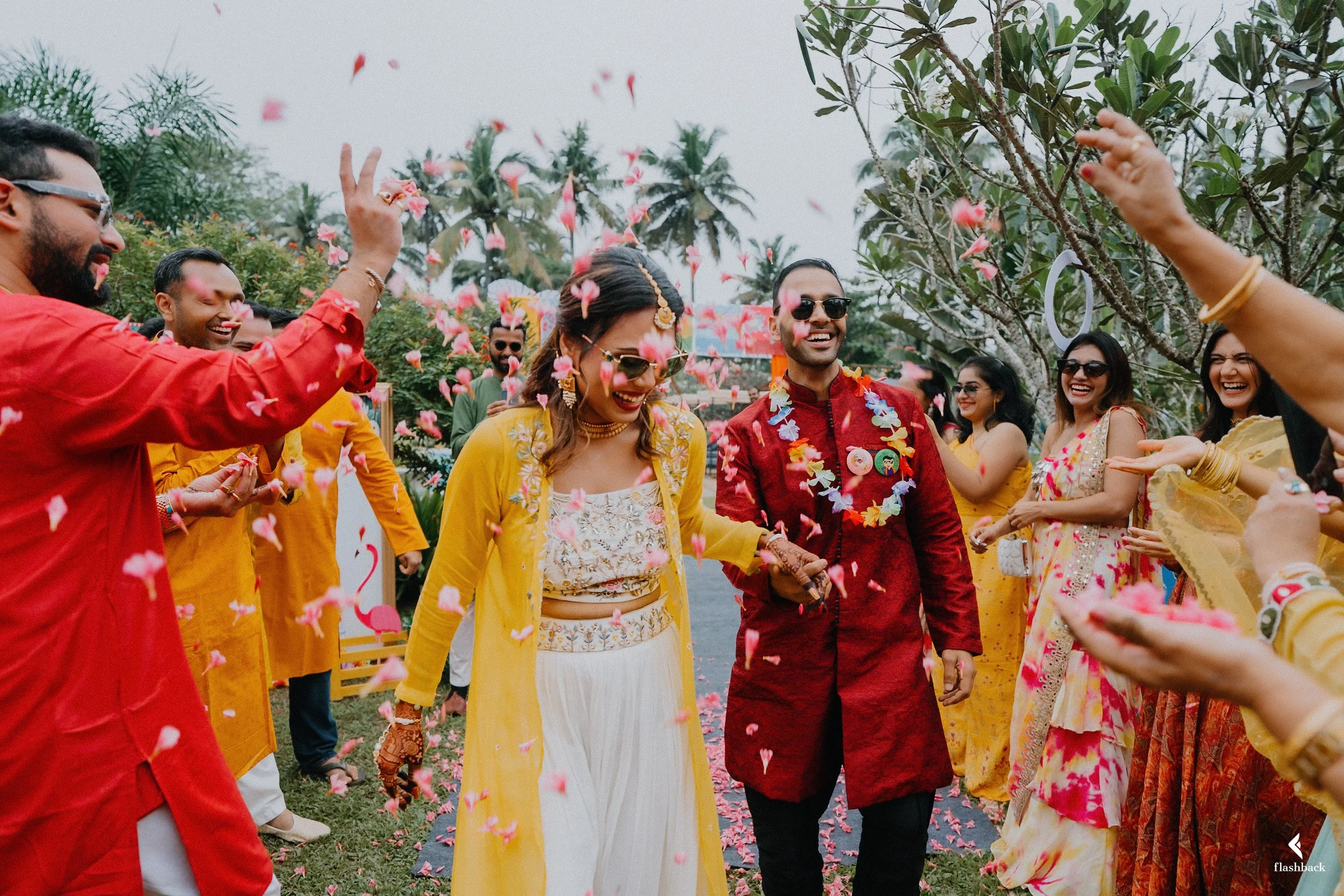 A group of people celebrating outdoors, dressed in colorful traditional clothing, with flower petals falling around them.