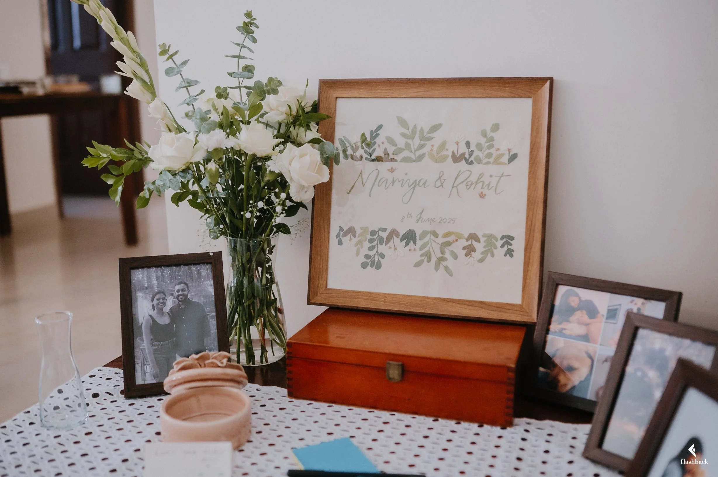 Decorative table with framed photographs, a glass vase with white flowers, a wooden box, and a framed sign that reads "Maringa & Rohit" with the date "8th June 2025".