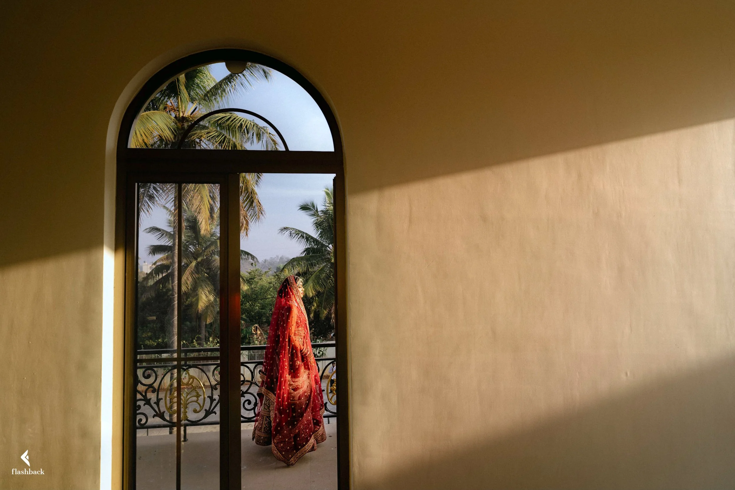 A woman dressed in traditional red Indian attire walking outside on a balcony with tropical palm trees in the background.