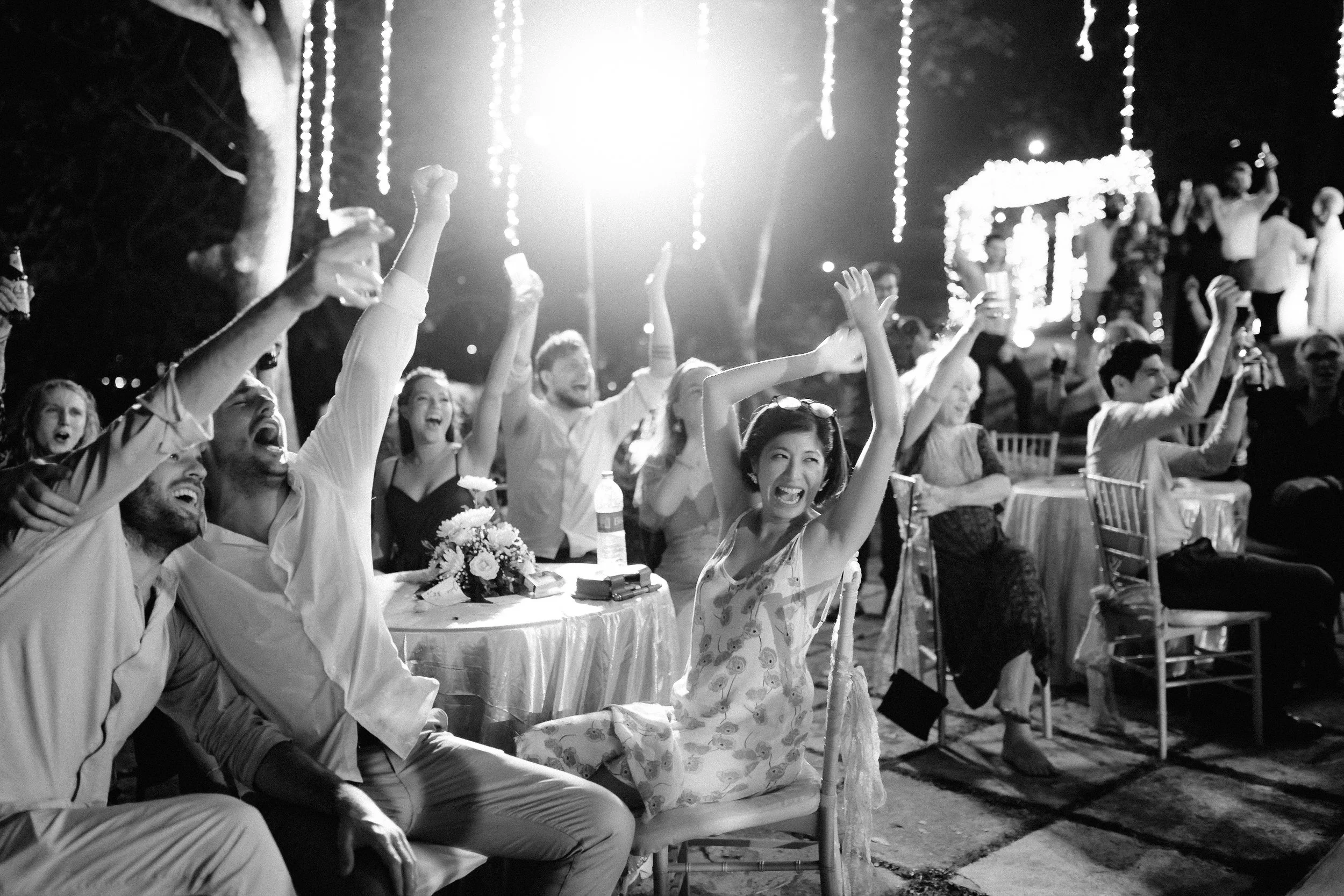 People at a nighttime outdoor celebration are cheering and raising their hands, with fairy lights hanging in the background and a decorated structure. Black and white photo.