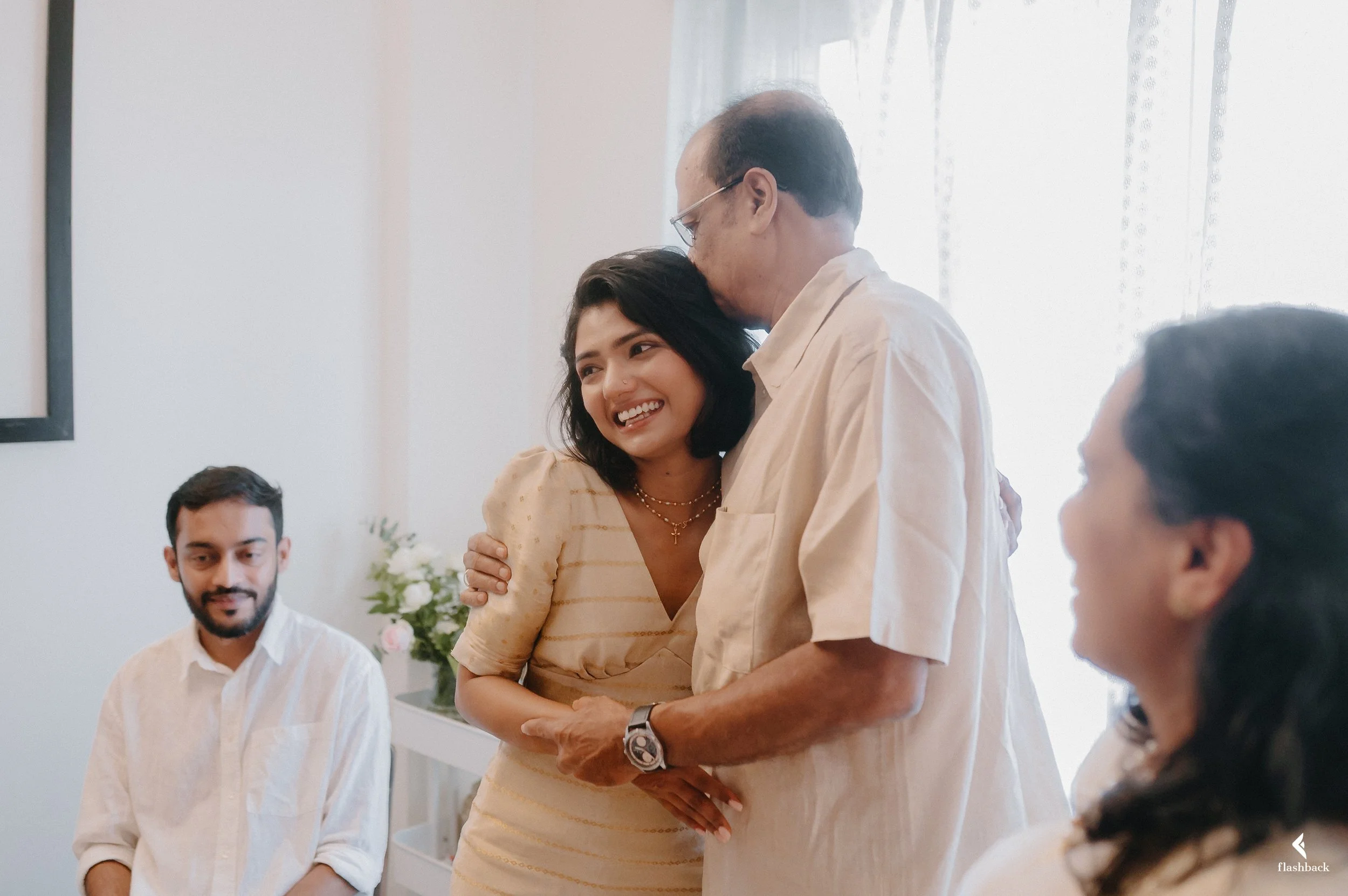 A joyful young woman hugging an older man in a white shirt at a family gathering, with two other family members, one man and one woman, sitting at a table in a bright room with sheer curtains and a flower arrangement.