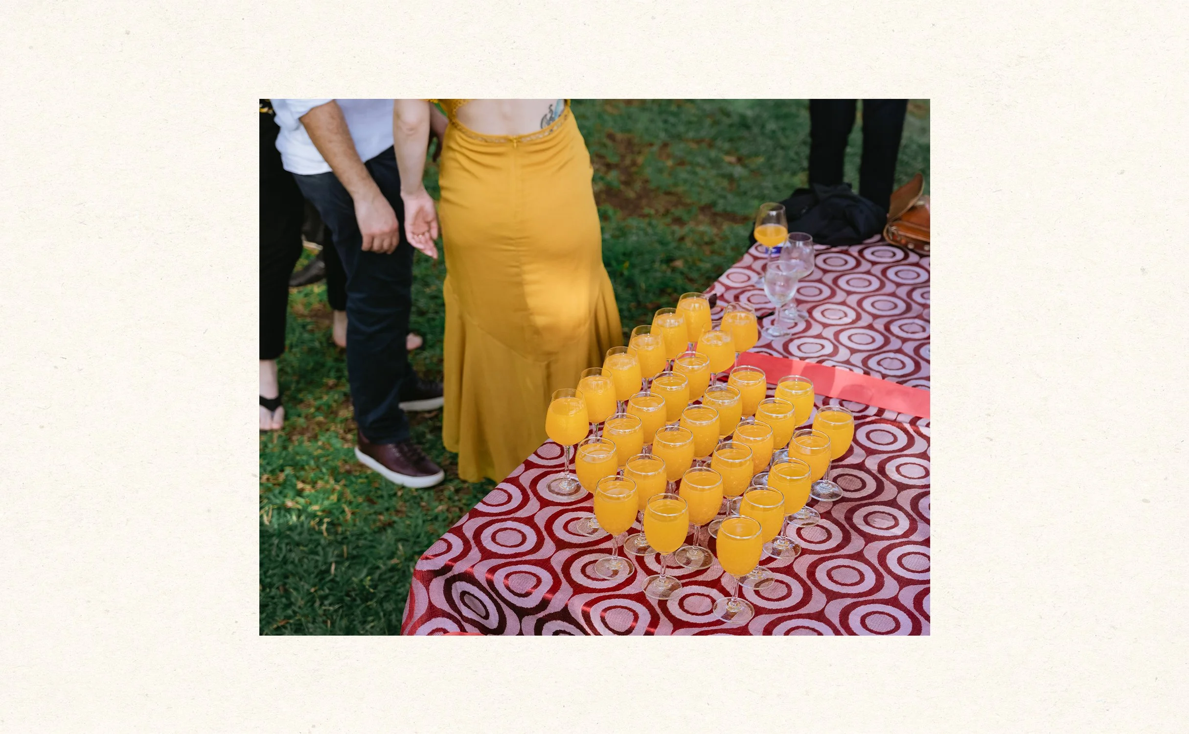 A table with many glasses of orange-colored drinks at an outdoor event, with people standing nearby.