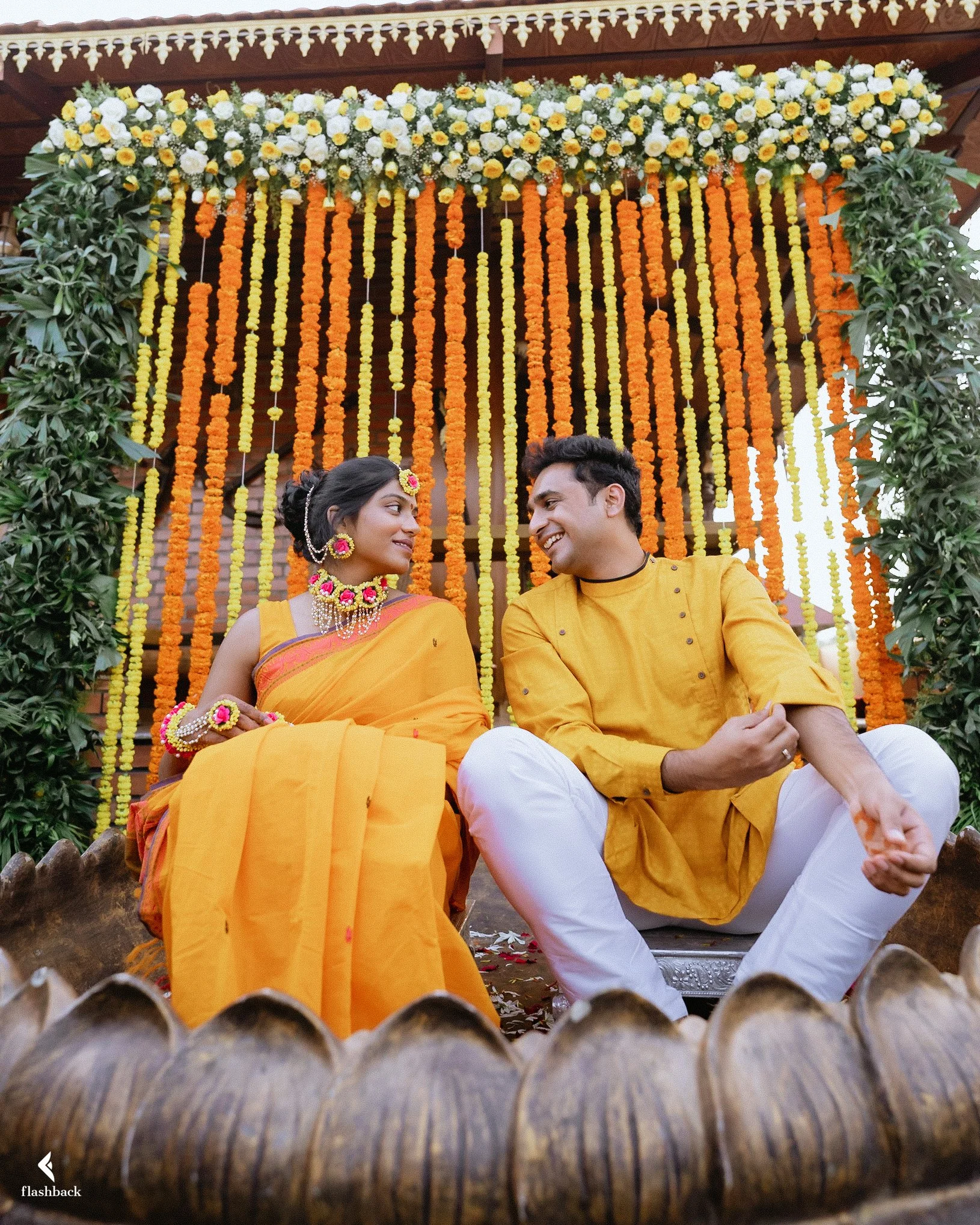 A couple dressed in traditional Indian attire sitting together during a celebration, with a yellow and orange floral backdrop.
