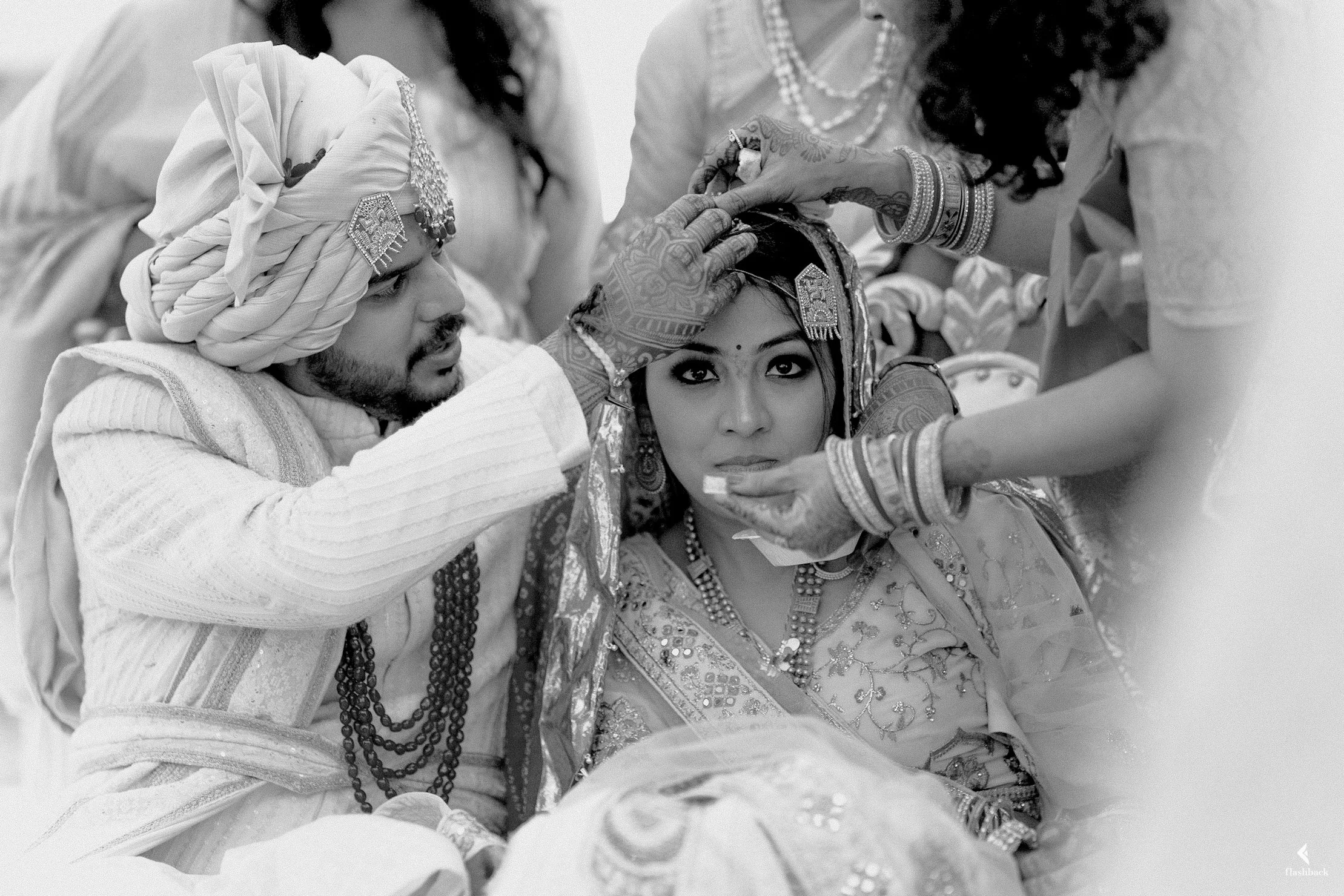 A traditional Indian wedding ceremony where a groom, wearing a turban and embellished clothing, is performing a ritual on the bride, dressed in ornate attire with jewelry. An elder woman assists in the ritual, while the bride looks directly at the ca