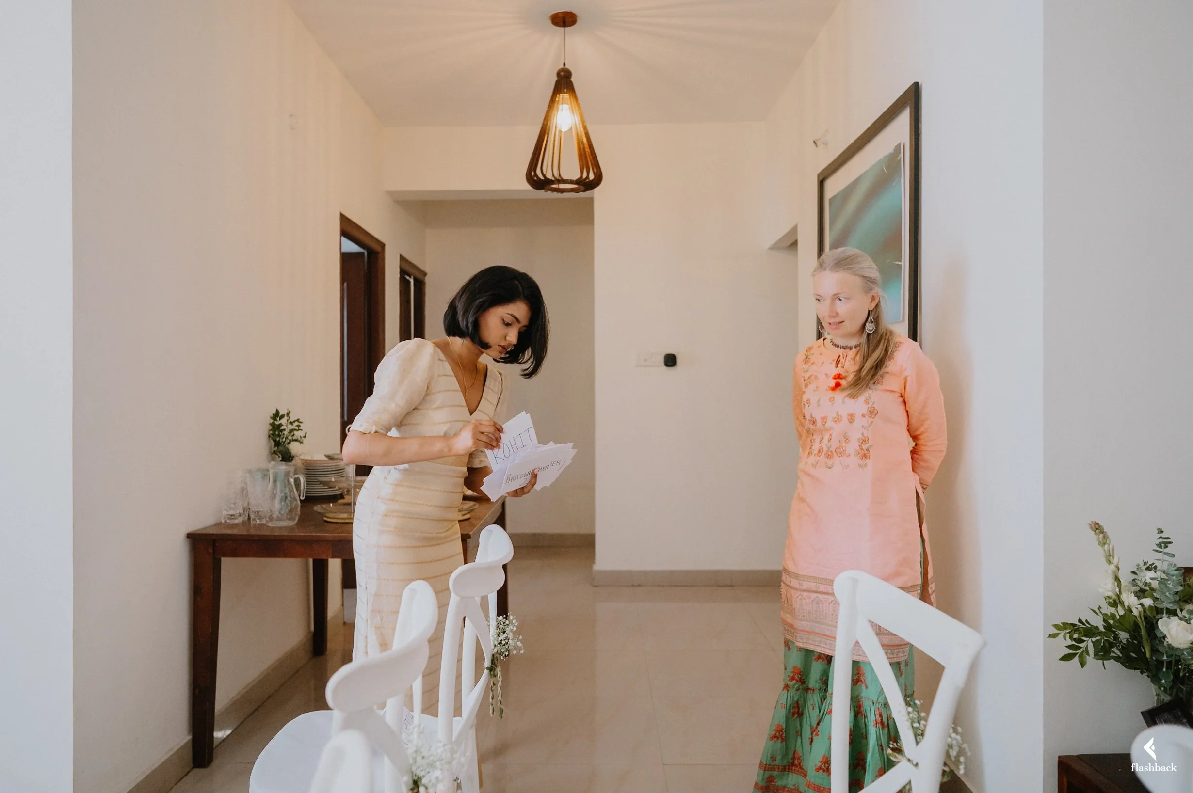 Two women in traditional Indian clothing standing in a room with white walls. One woman on the left is holding and looking at papers, and the woman on the right is standing with her hands behind her back, smiling. There are chairs with white covers a