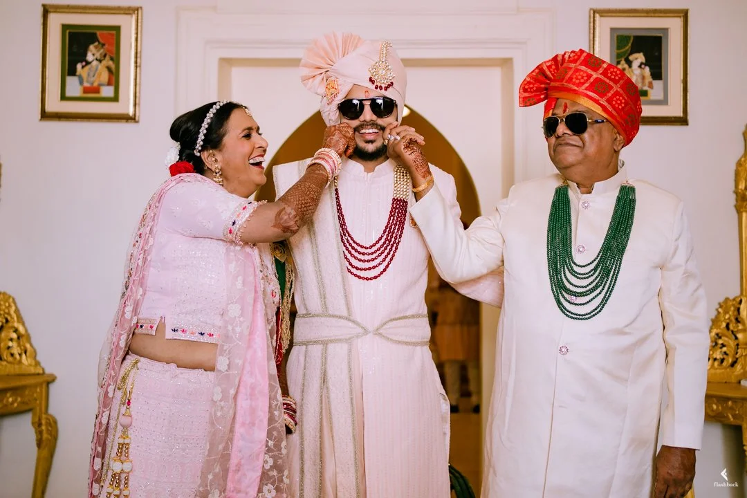 Indian wedding ceremony with three people dressed in traditional attire, two men wearing turbans and sunglasses, one woman applying henna on the groom's hand, all smiling and standing in a decorated room.