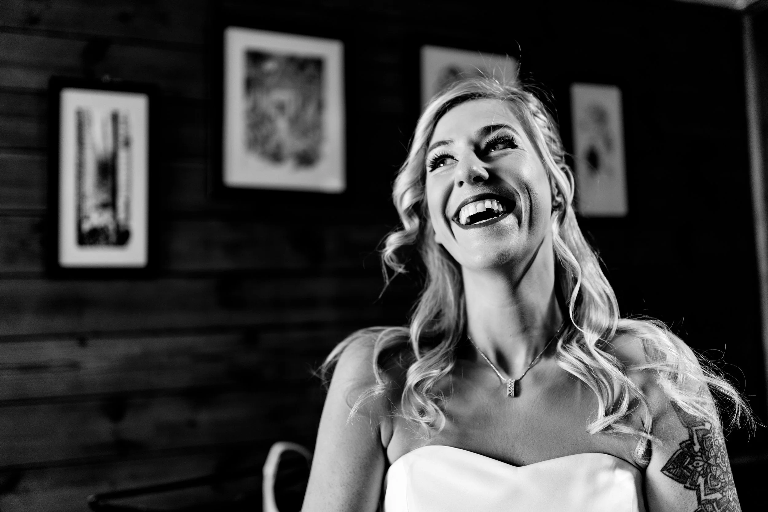 A woman with long, wavy blonde hair smiling and looking upwards, wearing a strapless top and a necklace, with framed artwork on a wooden wall in the background.