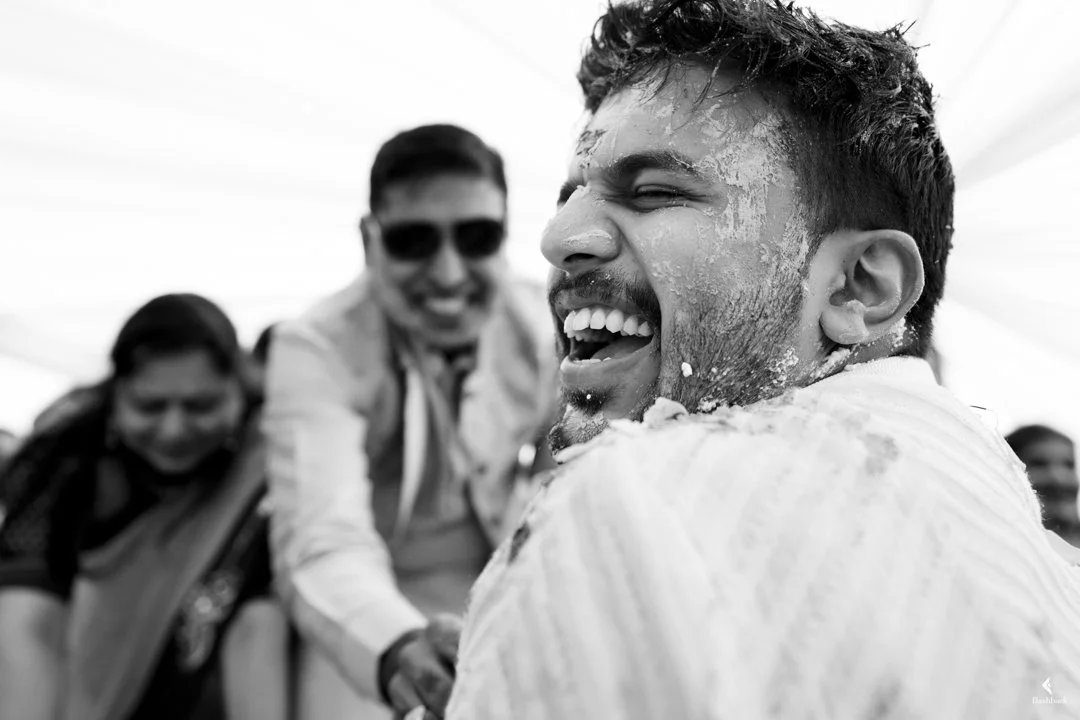 Three friends smiling and laughing, with one person in the foreground covered in cake or frosting, outdoors under a bright sky.