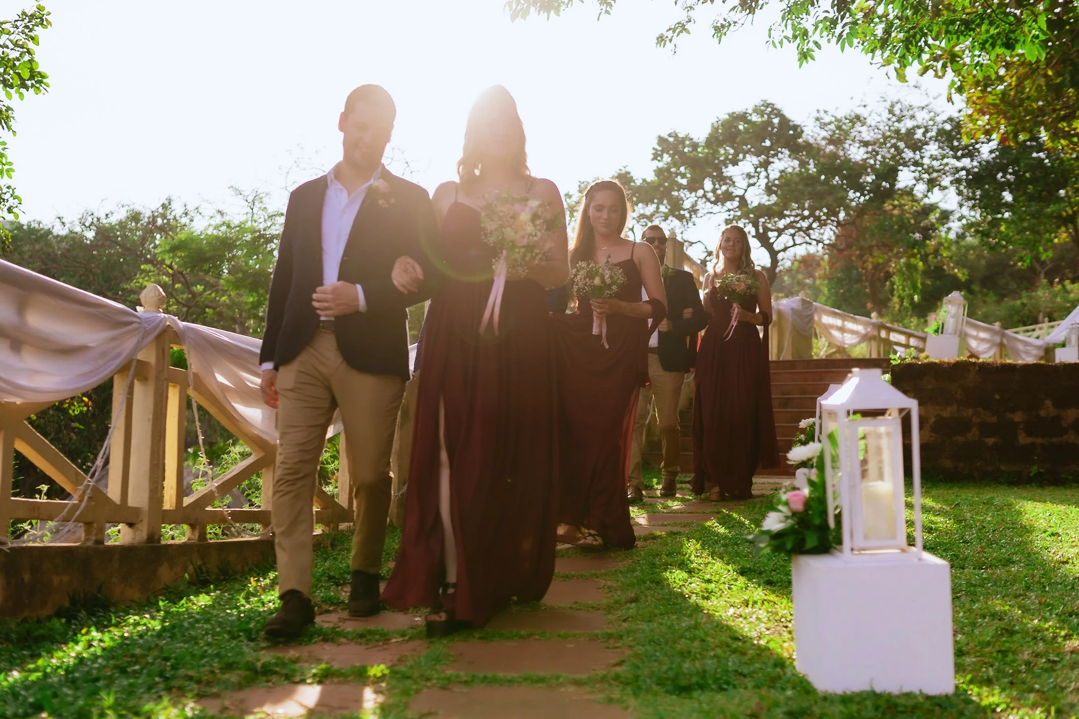 Wedding procession walking down outdoor garden steps with lanterns and floral decorations, sunlight shining through trees in the background.