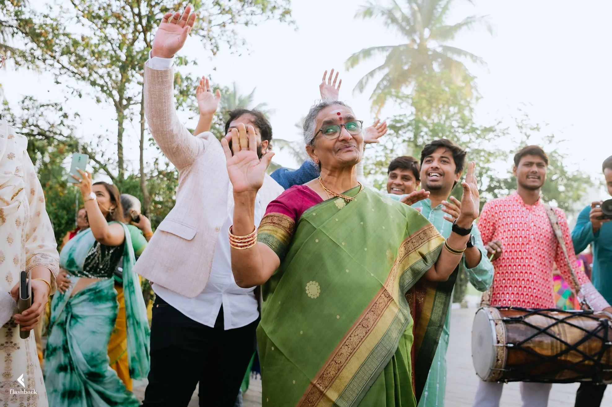 An elderly woman in a green sari waving and smiling at a festive outdoor gathering, surrounded by smiling people, some taking photos and playing musical instruments, with trees in the background.