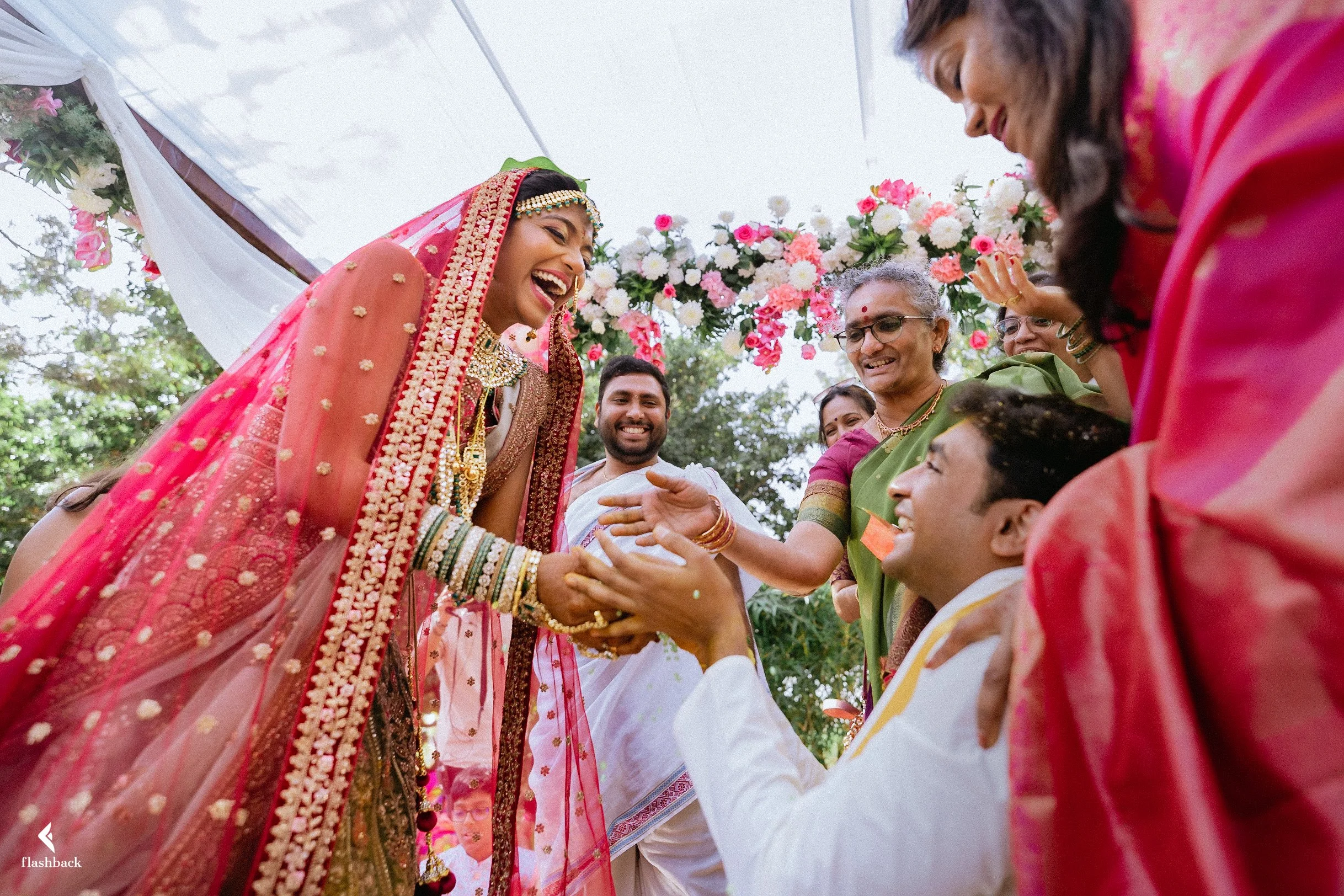 Indian bride in traditional red and gold wedding attire smiling and interacting with guests during a wedding ceremony with floral decorations and family members.