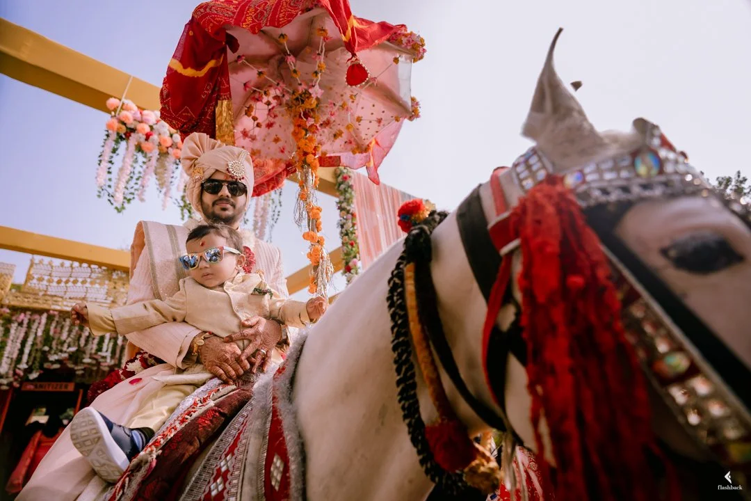 Man and child dressed in traditional Indian wedding attire, riding a decorated white horse with ornate adornments, under a decorative umbrella, during a festive celebration.