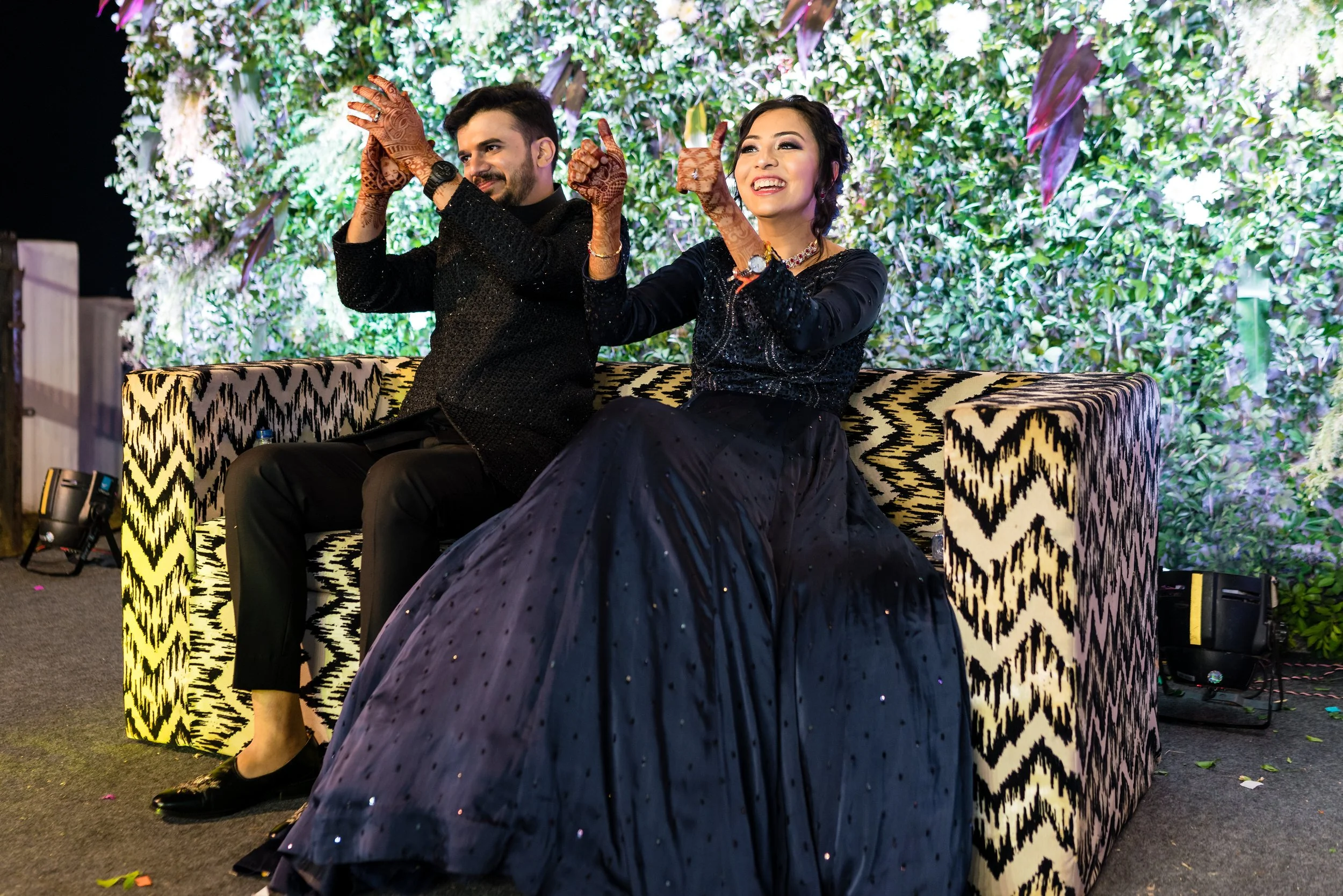 A man and woman sitting on a patterned sofa, smiling and raising their hands in a celebratory gesture. The woman is wearing a long, black, sparkly dress, and the man is wearing a black outfit with a textured jacket. They are in front of a lush, green