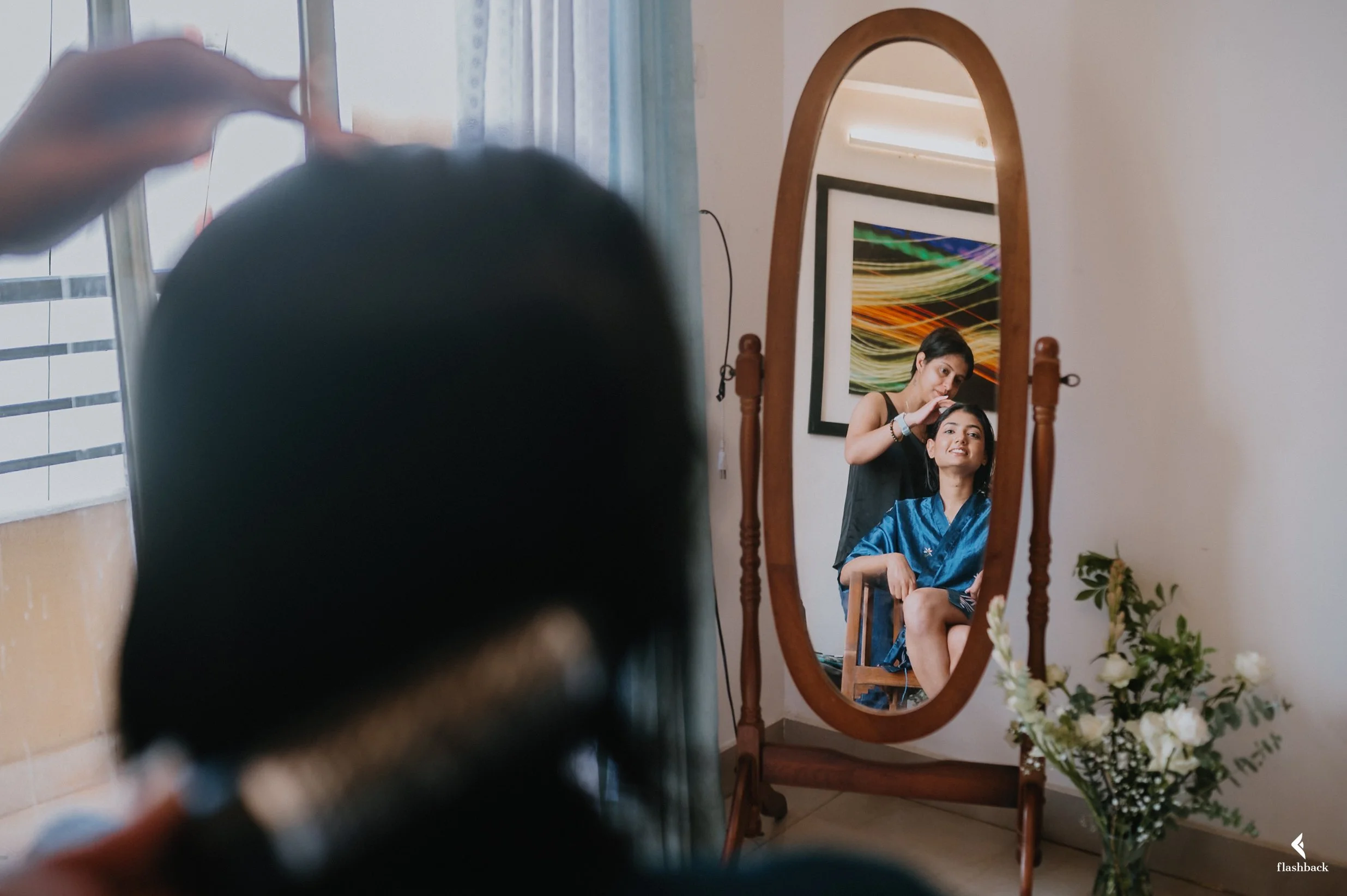 A woman getting her hair styled by a hairstylist, reflected in a full-length mirror, with the hairstylist standing behind her and adjusting her hair.