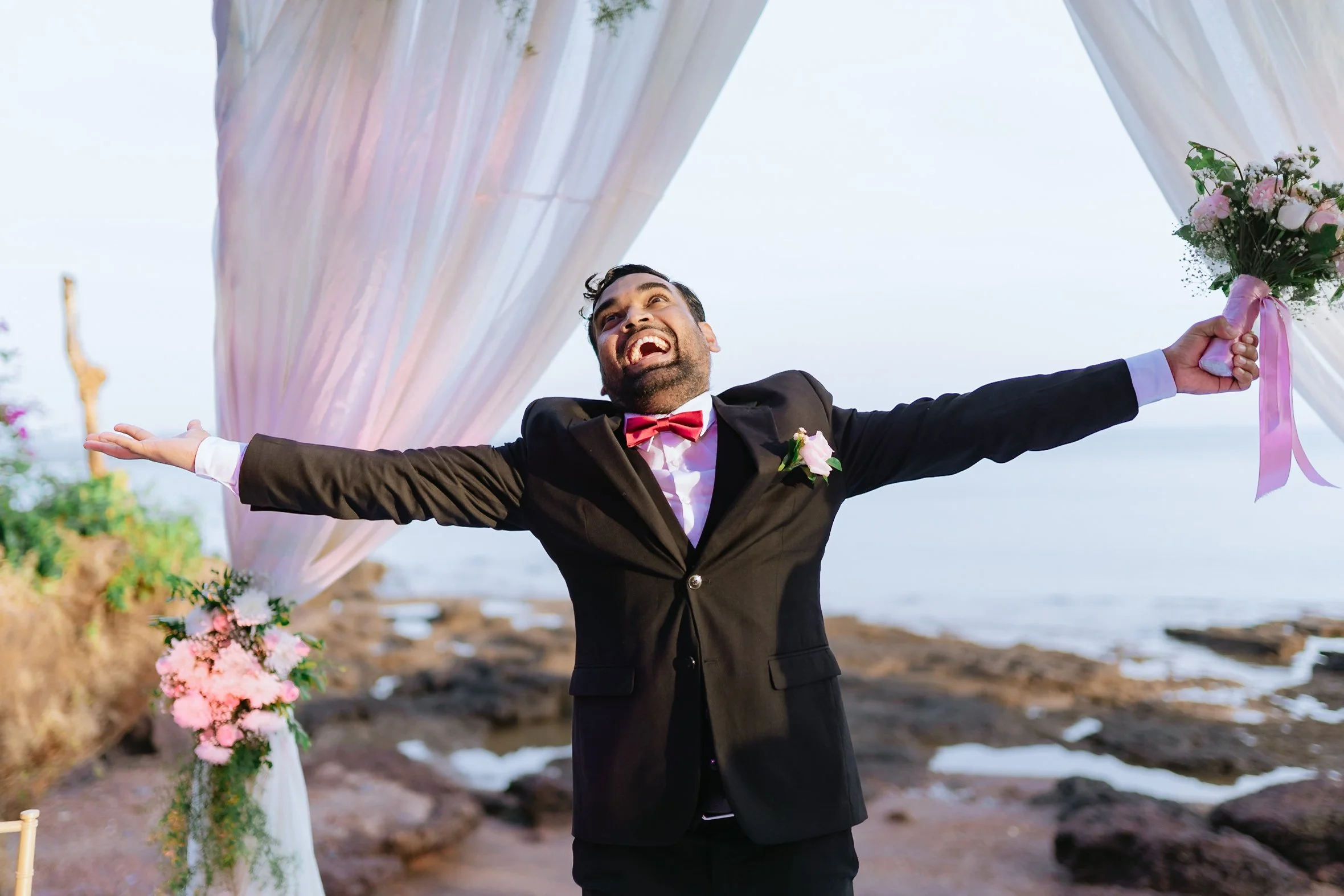 A man in a black suit with a pink bowtie and boutonniere celebrating at a wedding outdoors near the ocean, standing under a decorated archway with flowers and sheer fabric.