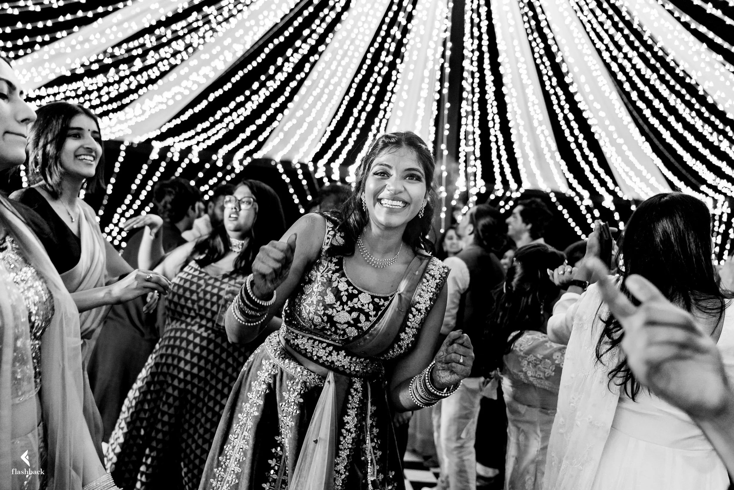 Woman in traditional Indian attire dancing and smiling at a celebration under a canopy of string lights, with other guests around her.
